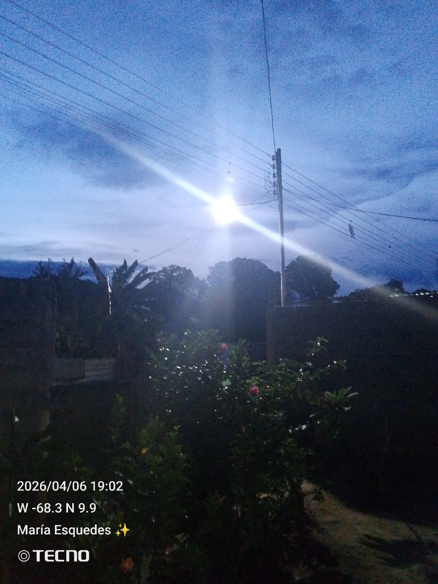The image shows an outdoor scene at night in Tinaquillo, Venezuela, with the timestamp indicating April 6, 2026, at 19:02. The coordinates are W -68.3 N 9.9. A bright light source dominates the center of the frame, likely a street lamp. There is a utility pole with wires overhead, obscured by the light. Vegetation, including bushes and trees, is visible in the foreground and background. A wall or roofline appears in the middle ground on the right side. The sky is a dark blue with some cloud cover. The photographer is identified as Maria Es quedes. A TECNO watermark is visible at the bottom.