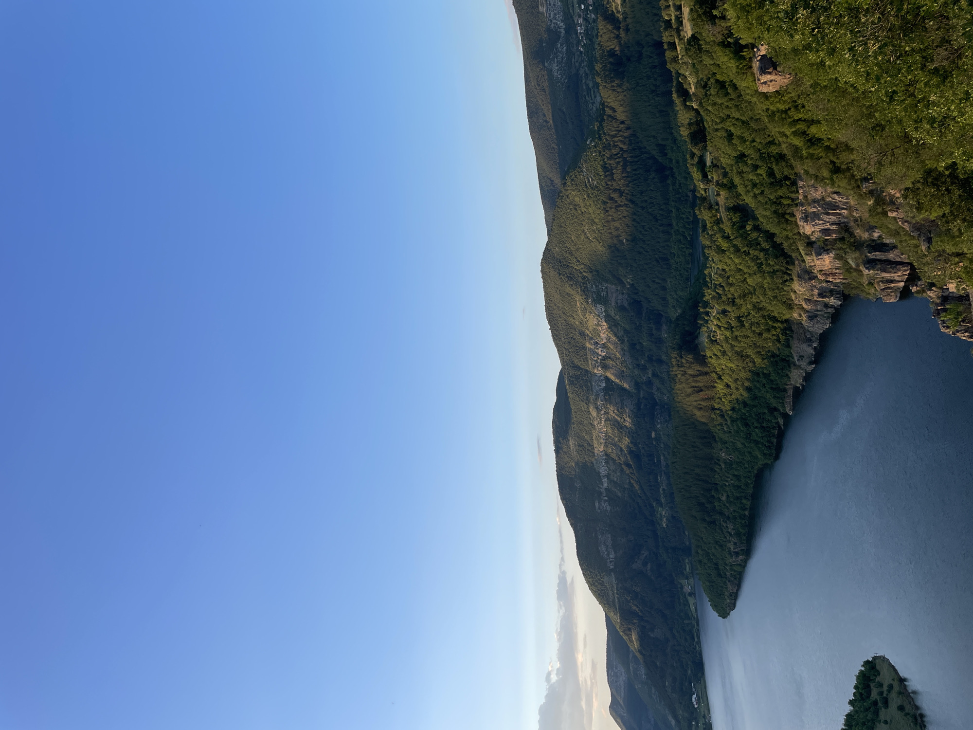A vertical, high-angle shot captures a vast, tranquil body of dark blue water, likely a reservoir, at the base of a verdant, mountainous landscape. The water occupies the lower left and center of the frame, with a narrow inlet extending towards the bottom left corner, bordered by a small, grassy landmass dotted with trees. Steep, rocky cliffs, some with visible strata and vegetation, rise from the water's edge on the right side, transitioning into densely forested slopes. The mountains, covered in a thick carpet of evergreen trees, are bathed in the soft, golden light of either sunrise or sunset, creating long shadows and highlighting the contours of the terrain. The sky above is a clear, expansive gradient of pale blue, transitioning to a deeper hue towards the top of the frame, with only a few wispy clouds visible near the horizon on the left. In the distance, the peaks of the mountains recede, showcasing layers of forested slopes. The overall scene is one of serene natural beauty, evoking a sense of peaceful isolation. The location is identified as the area around Borovitsa, Bulgaria, suggesting a rural or natural setting. No people or specific man-made structures are prominently visible, emphasizing the untouched, natural environment.