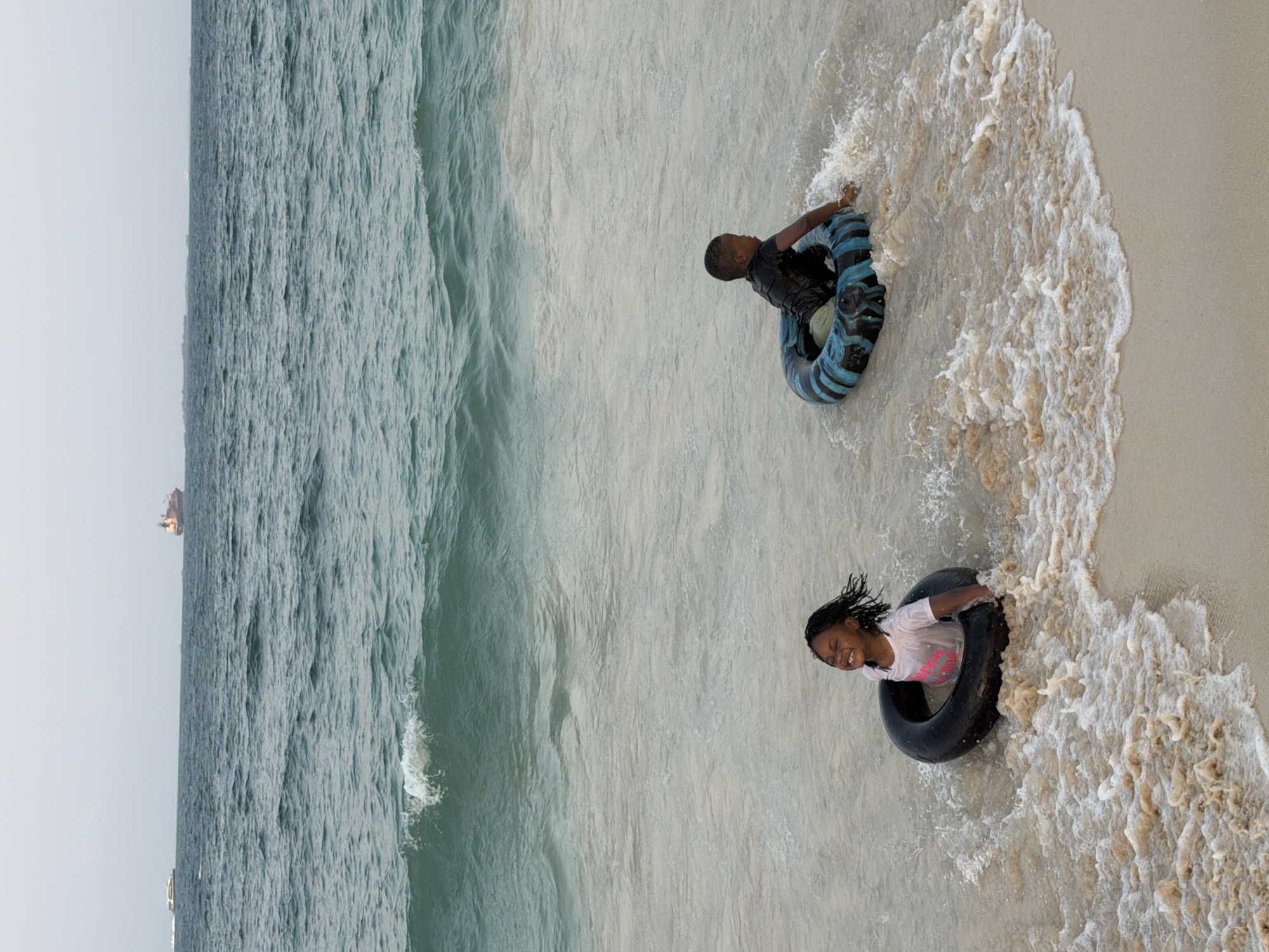 Two young children, a girl and a boy, are playing in the shallow waters of the ocean on a sandy beach. The girl, on the left, is wearing a white t-shirt and is positioned inside a black inner tube, with her face turned upwards and a wide smile. Her arms are resting on the edge of the tube. The boy, on the right, is also inside a black and blue striped inner tube, wearing a black t-shirt. He is facing away from the camera, looking towards the sea, with his hands on the tube. Gentle waves are washing around the children. In the background, the ocean extends to the horizon under a hazy sky. Two ships are visible on the water, one further out and a smaller vessel closer to the left side of the frame. The scene depicts a recreational activity at a beach location, likely in Dar es Salaam, Tanzania, given the presence of ships on the Indian Ocean and the typical beach environment.