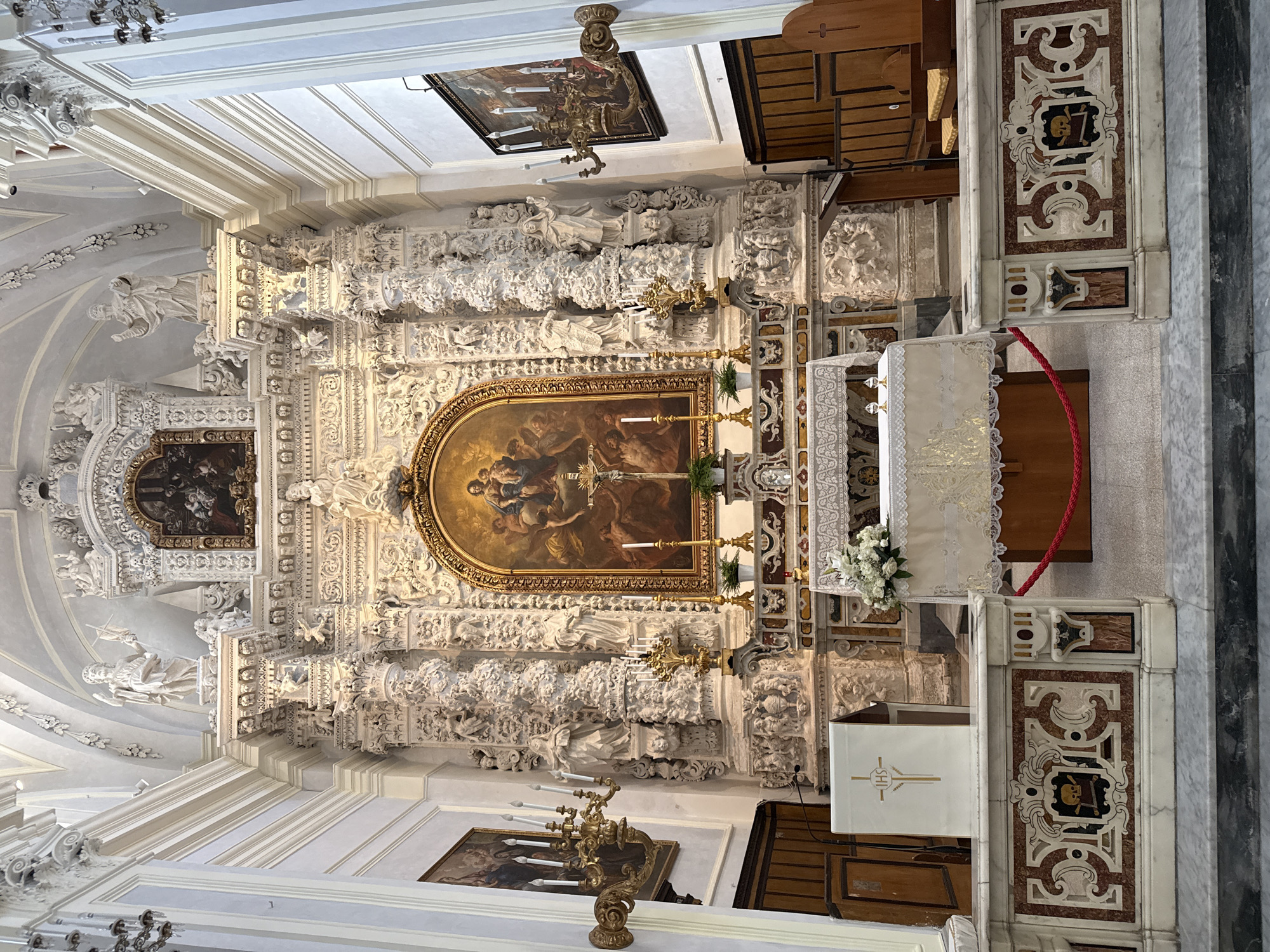 The image displays the ornate interior of a church, specifically focusing on the main altar and its surrounding apse. The style is characteristic of Baroque or Rococo architecture, featuring an abundance of intricate white stucco decorations.
At the heart of the scene is a large altarpiece painting, housed within a magnificent gilded frame. The artwork depicts a religious narrative, likely centered on the Virgin Mary and Christ, surrounded by other figures such as angels or saints, rendered with dramatic lighting and warm colors. Flanking the painting are two prominent golden candelabras, each holding multiple candles. On the altar table below, a white cloth with delicate lace trim is adorned with a vase of fresh white flowers, possibly lilies or roses, adding a touch of serene beauty.
The walls surrounding the altarpiece are a spectacle of detailed white stucco work. This includes sculpted figures of saints or angels emerging from clouds, intertwined with rich floral motifs, garlands, cherubs, and complex architectural elements like columns, pilasters, and cornices. The ceiling is vaulted and also features elaborate white ornamentation.
To the right of the altar, a simple white stand or lectern is visible, marked with the "IHS" monogram. Adjacent to this, decorative marble panels in shades of white, brown, and black display geometric and scroll patterns. One panel features a distinct central motif that appears to be a skull and crossbones, a classic memento mori symbol. A red rope barrier is also present on the right side. Overhead, chandelier-like light fixtures hang from the ceiling, and other paintings can be glimpsed on upper walls.
The bright illumination suggests daylight. No individuals are present, allowing the viewer to absorb the intricate artistry of the sacred space. This grand and historically rich setting is located in Monopoli, Italy.