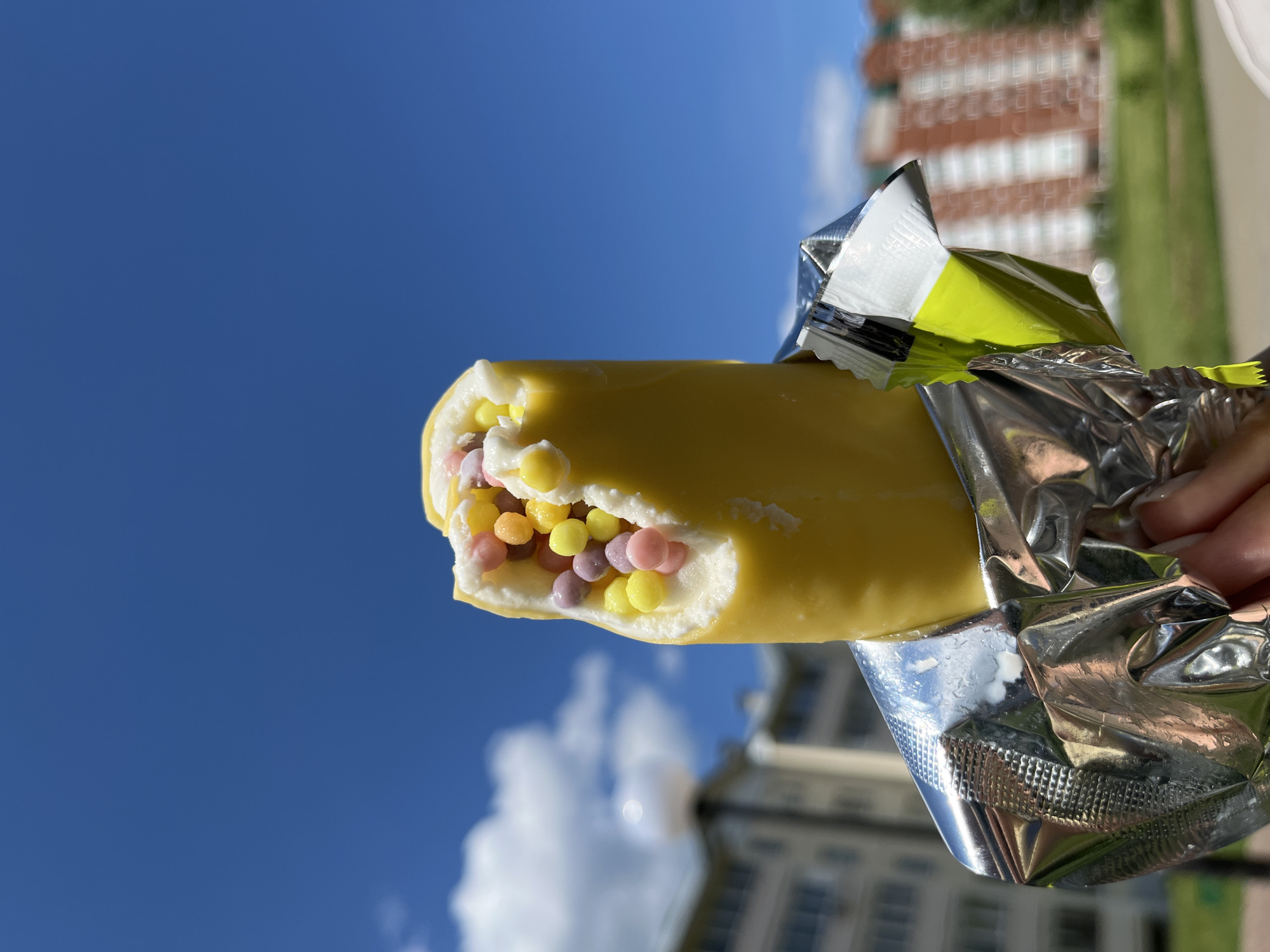 A hand holds a partially consumed, yellow-coated ice cream bar, unwrapped from its crinkled silver foil packaging, against a bright blue sky and a blurred urban background in Chaikovsky, Russia. The central subject is an ice cream bar, approximately two-thirds visible, with a smooth yellow outer layer. A distinct bite has been taken from the top, revealing a white, creamy interior densely packed with small, spherical candies in pastel shades of pink, yellow, purple, and orange. The reflective silver foil wrapper shows visible crinkling and what appears to be melted ice cream residue or condensation droplets. A section of a yellow-green inner lining is visible on the top right edge of the wrapper. A light-skinned hand, with fingers partially visible, grips the foil at the bottom-center of the frame.
The background is characterized by a clear, vibrant blue sky filling the upper portion of the image, featuring a few soft, white cumulus clouds on the left. Below the sky, blurred multi-story residential buildings are discernible. On the left, light-colored buildings with multiple windows are visible, along with a white, spherical street lamp on a pole. On the right, a more distant building with reddish-brown vertical architectural elements is partially visible. Green foliage, likely grass and trees, is present in the blurred mid-ground and foreground, indicating an outdoor, daytime setting within a landscaped residential or park area. The scene is illuminated by direct sunlight, casting reflections on the ice cream wrapper.