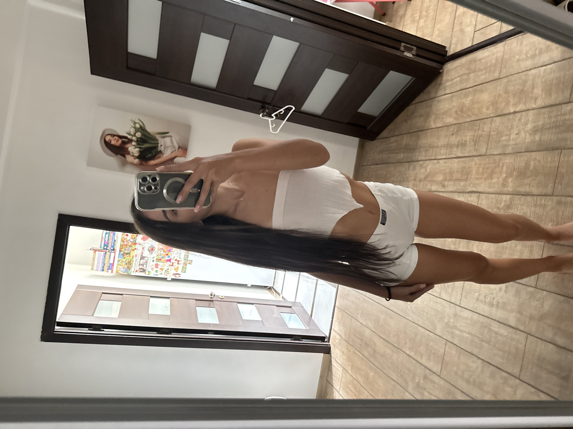 A woman is taking a mirror selfie in a room with light brown tiled flooring and two wooden doors. The woman is wearing a white strapless top and white shorts. She has long dark hair. To her left, a door is slightly ajar, revealing a glimpse of another room with posters on the wall. To her right, a dark wooden door is closed, with a white hanger hanging on its knob. In the background, on the wall, a framed picture of a woman holding tulips is visible. The room appears to be in a residential setting, possibly an apartment in Chișinău, Moldova, given the context.