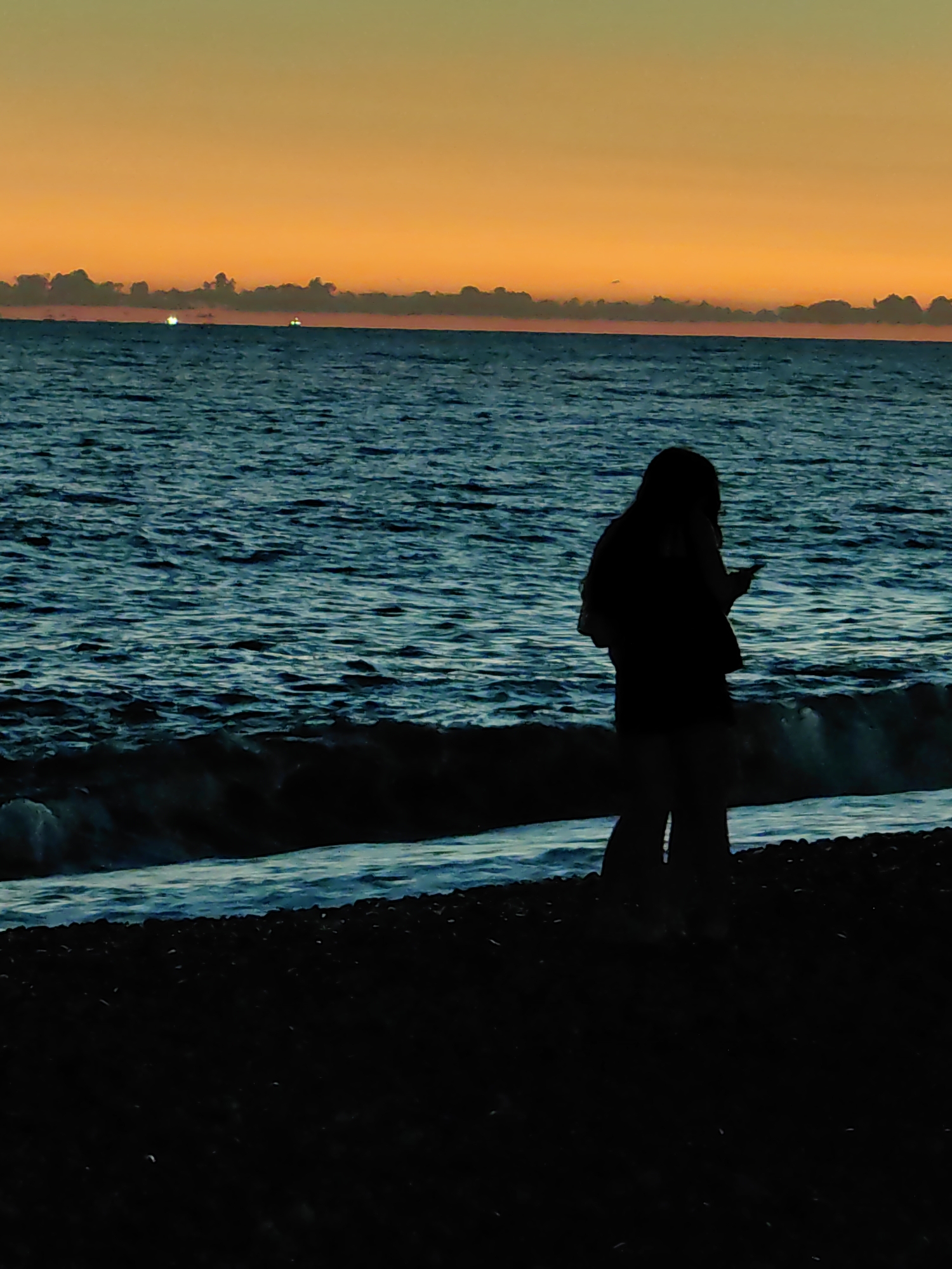 This is a silhouette image taken at dusk on a beach in Sochi, Russia. The sky is a vibrant gradient of orange and yellow. In the foreground, two figures are standing on a dark, pebbly beach. The figure on the left is largely obscured by shadow, but appears to be a woman with long hair, holding a phone in her right hand. The figure on the right is a partial silhouette of what appears to be another person standing next to the first. The sea is a deep blue, with visible waves breaking on the shore. Two distant lights are visible on the horizon, possibly from boats. The overall atmosphere is serene and twilight.
