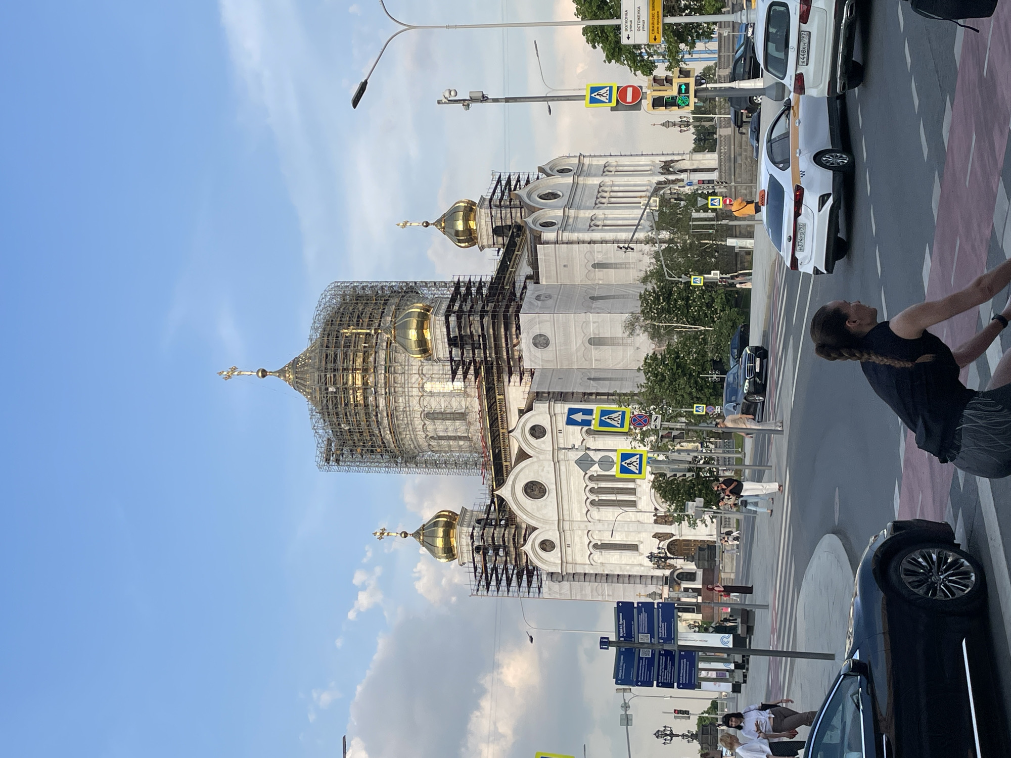 A street-level, medium shot depicts the Cathedral of Christ the Savior in Moscow, Russia, undergoing construction or renovation. Scaffolding surrounds the largest dome, partially obscuring its gold surface. Adjacent domes are also visible. The cathedral is white and ornate. The photo captures a bustling city scene. In the foreground, multiple parked cars line the right side of the street. Pedestrians cross the street at a crosswalk marked by blue and yellow signs. Street signs with Cyrillic text are mounted on poles. Traffic lights hang overhead. The sky is blue with scattered white clouds. The overall lighting suggests it's daytime.