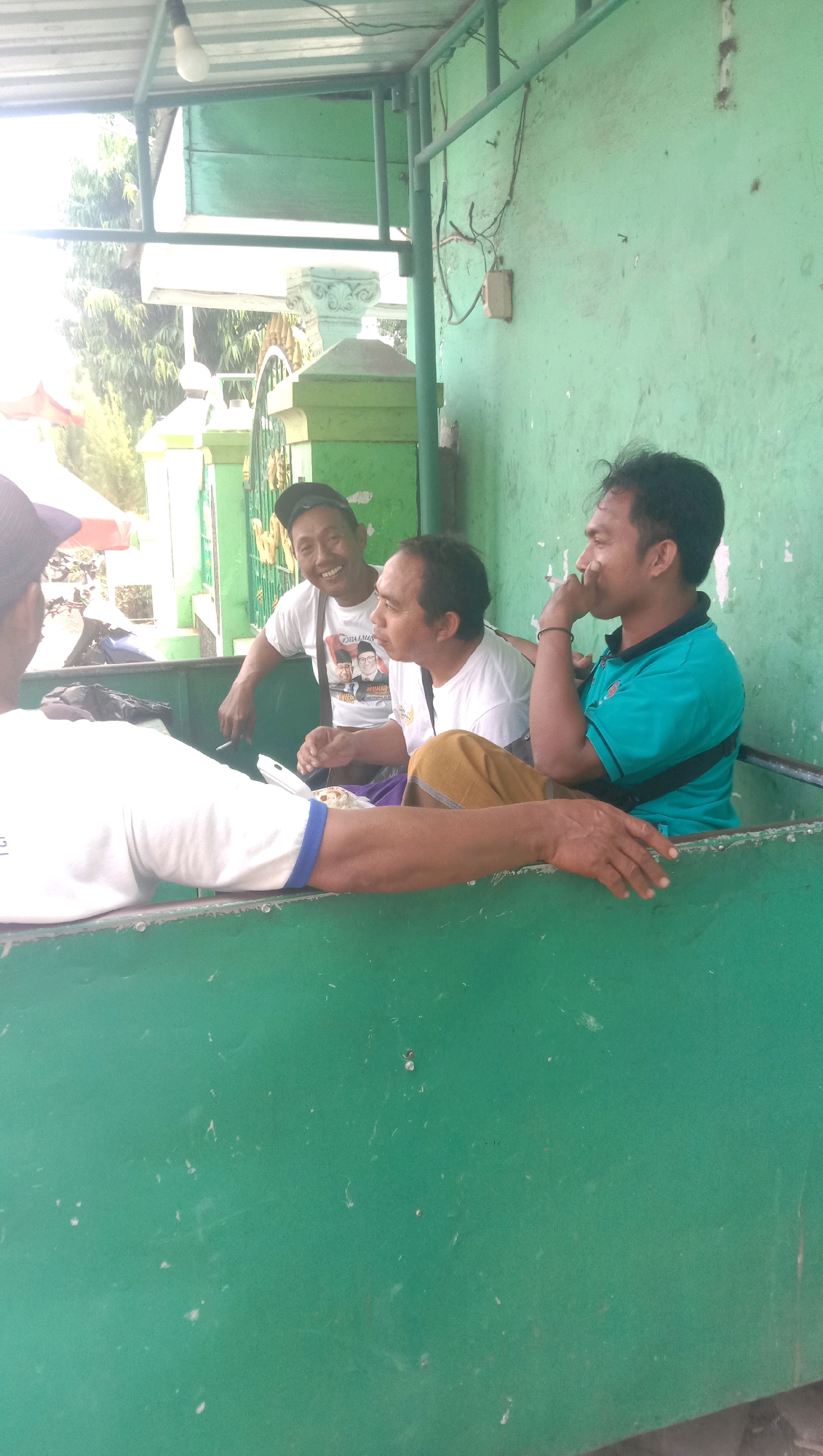 The image depicts an outdoor scene in Karang Anyar, Indonesia, featuring four individuals interacting in a sheltered area. The environment appears to be rustic, with a corrugated metal roof supported by green metal beams. The background shows foliage and what looks like a fence or gate.
In the foreground, three men are seated in a green, open-topped container, possibly a truck bed or a makeshift seating area. One man, on the left, is partially visible and appears to be wearing a white t-shirt. The middle man, wearing a white t-shirt with a graphic of three men on it, is smiling and holding a cigarette. To his right, another man in a turquoise t-shirt is looking away and has his hand near his mouth, suggesting he might be smoking or contemplating. The fourth person is only partially visible in the lower left corner, wearing a white shirt with blue trim. The men seem to be engaged in a casual conversation or a shared moment, indicated by their relaxed postures and expressions.
The overall impression is of a common gathering spot, perhaps a rest stop or a gathering place for locals in a rural or semi-urban area of Karang Anyar. The green color scheme of the structure is prominent, and the lighting suggests it is daytime.