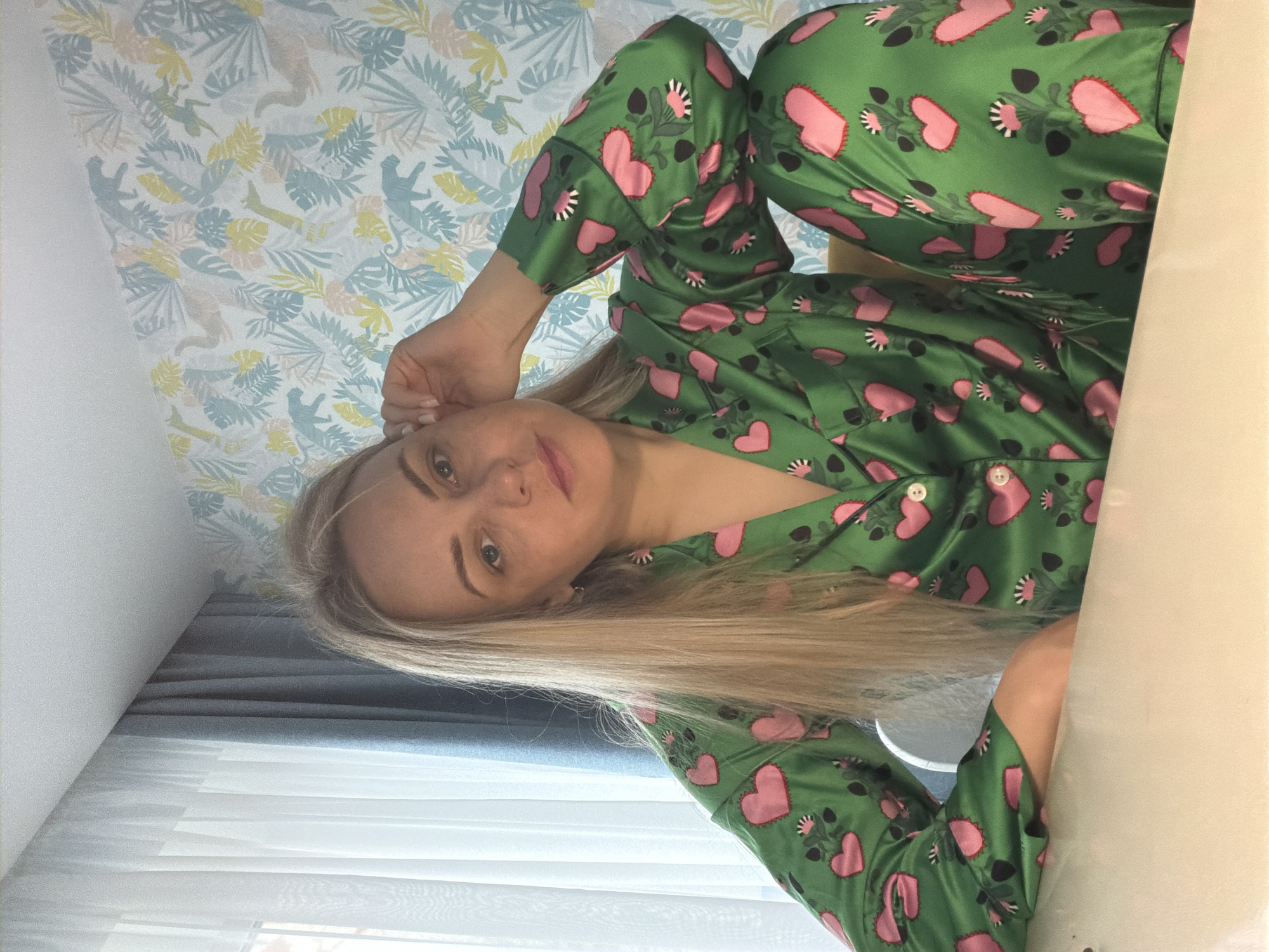 An individual with long, straight blonde hair and fair skin is captured in a close-up indoor shot, looking directly at the camera with a neutral expression. She is wearing green satin pajamas adorned with large pink hearts and smaller black and white floral or leaf patterns. Her left hand is raised towards her head, near her ear.
The setting appears to be a room, possibly a bedroom or living area, in Votkinsk, Russia. The background features a distinctive patterned wallpaper with light blue, yellow, and beige botanical elements, including leaves and stylized animal figures resembling monkeys or lemurs, set against a light background. A section of plain white wall is also visible. To the left and below the person's head, window blinds, likely white or light gray, are visible, partially covered by blue-grey curtains. A light-colored surface, possibly a table or desk, partially obscures the lower right side of the frame. The lighting suggests it is daytime. There is no visible text in the image.