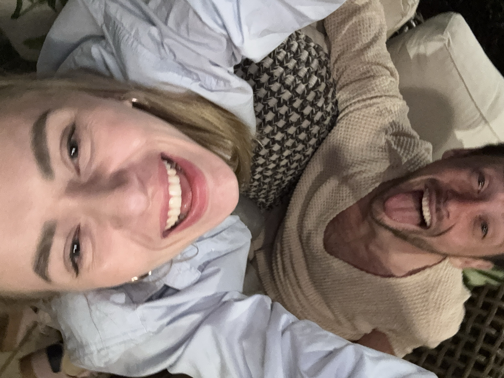 A selfie shows a woman and a man, both smiling broadly and looking towards the camera. The woman is on the left, wearing a light blue shirt. The man is on the right, sticking his tongue out, and wearing a beige sweater. They appear to be lying down, possibly on an outdoor seating area, with a patterned pillow between them. The background suggests they are in an outdoor location with furniture and potentially some foliage.