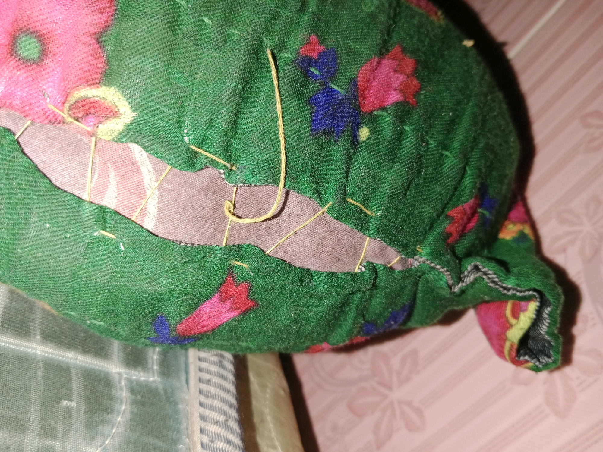 The image shows a close-up of a green fabric item with a floral pattern, possibly a cushion or bag, that has been torn and is being repaired with yellow thread. The fabric is gathered and bunched in places, indicating it is likely a soft item. There is a rip or tear exposing a patterned inner layer of fabric, with visible stitches holding the green fabric together around the opening. The background is a blurry pink surface with a subtle floral design. The image does not contain any discernible elements that would place it specifically in Ringinrejo, Indonesia, or provide any context related to that location. It is a still photograph of an object.