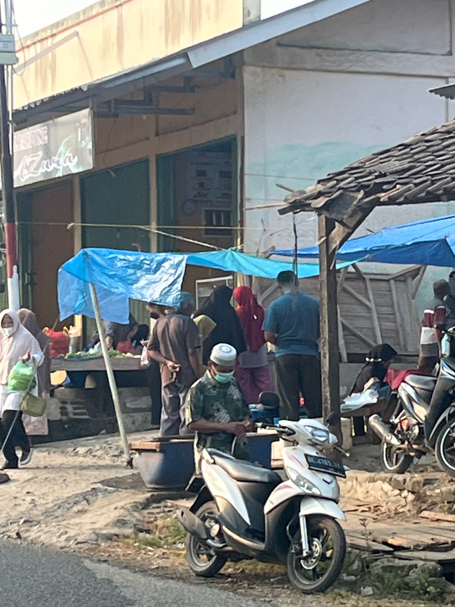 The image depicts a street scene in Kebumen, Indonesia, featuring a marketplace or roadside stall. Several individuals are present, some wearing religious attire such as hijabs and peci hats. A blue tarp is extended over a makeshift stall, providing shade. Goods, likely produce, are displayed on a table. Two motorcycles are parked in the foreground, one white and the other black. The environment suggests a warm climate, with clear skies visible and sparse vegetation along the roadside. The buildings in the background appear modest, with signs indicating businesses. The overall impression is that of a typical day in a local Indonesian community.