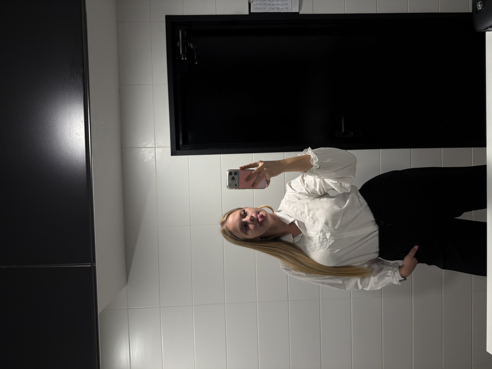 A young woman with blonde hair is taking a selfie in a bathroom with white tiled walls. She is wearing a white button-down shirt with lace detailing and black pants. She has one hand in her pocket and is sticking her tongue out at the camera. In the background, a black door is visible. There is no indication of the city of Izhevsk, Russia in the image.