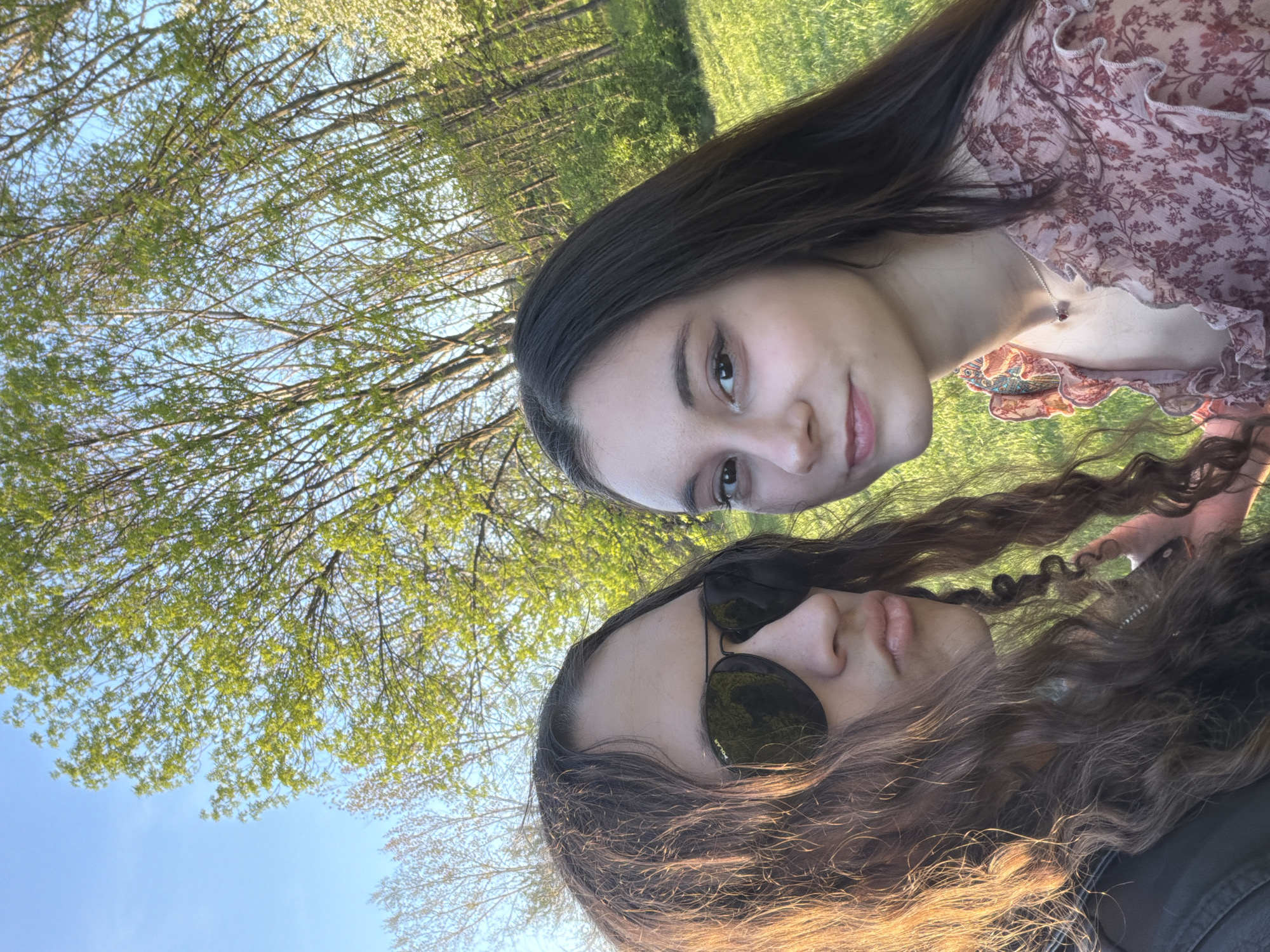 Two young women are captured in a close-up outdoor portrait, looking towards the viewer. The image is oriented sideways, but depicts them facing upwards against a backdrop of trees and sky.
One woman has long, dark brown, straight hair and is wearing a light pink floral top with ruffles around the neckline. She has a gentle smile and a silver necklace with a small, dark pendant. The second woman has long, dark brown, curly hair and is wearing dark sunglasses, with the word "POLAR" visible on the right lens. Her lips are tinted with light pink.
The setting is an outdoor natural environment, likely a park in Sofia, Bulgaria. The background features a clear blue sky and numerous trees with vibrant green leaves, suggesting spring or early summer foliage. Green grass is visible in the distance. The lighting is warm and golden, indicative of late afternoon or early evening sunshine, casting a pleasant glow on the subjects. The overall atmosphere is casual and relaxed.