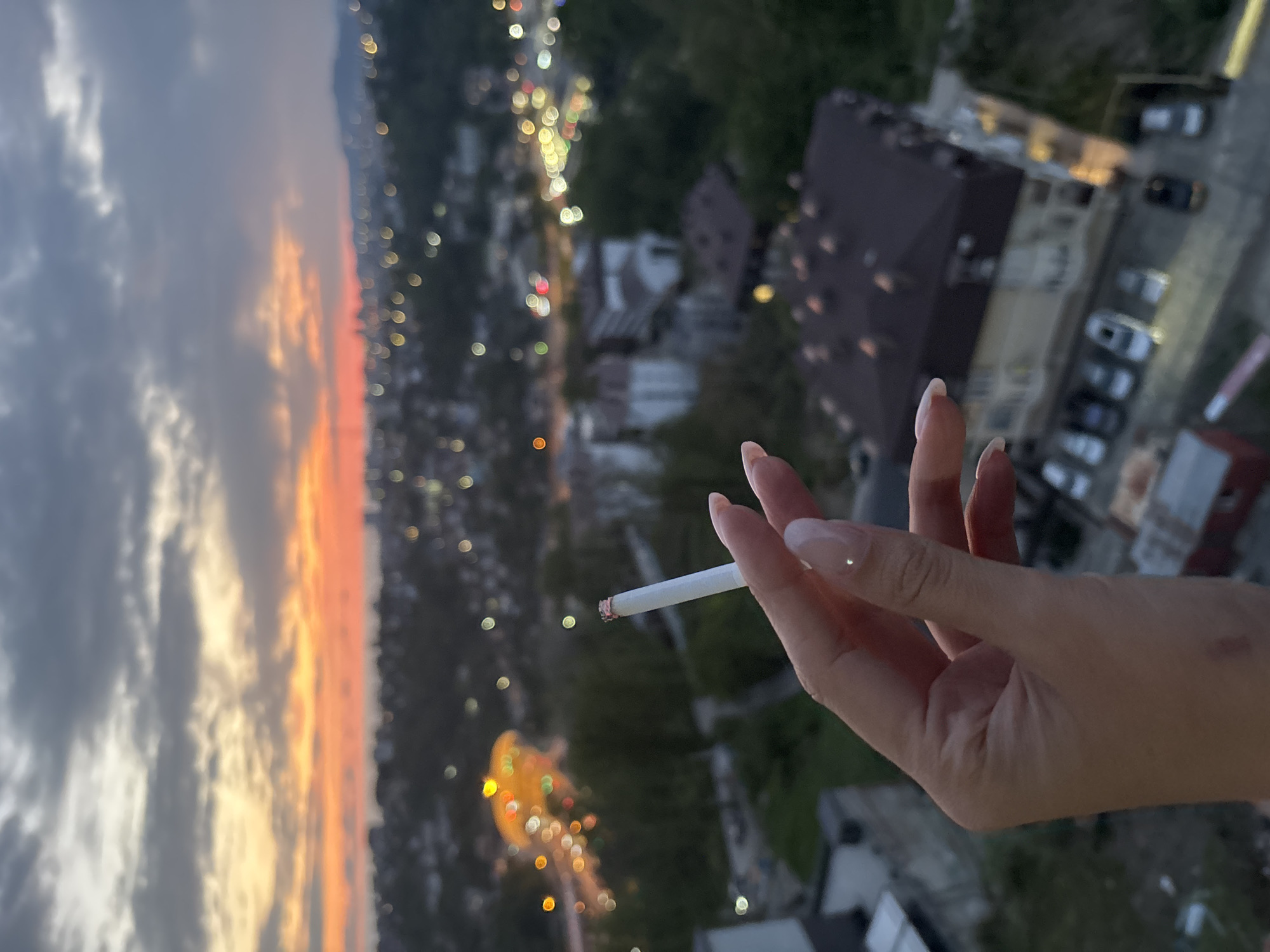 The image shows a hand holding a lit cigarette against a backdrop of a cityscape at dusk, likely in Sochi, Russia. The sunset is visible on the left with orange and yellow hues. The city stretches out with blurred lights and building silhouettes. A building with a dark roof is noticeable on the right. Cars are parked near a multi-story building. The hand is fair-skinned with manicured nails and is holding the cigarette between the index and middle fingers. The end of the cigarette is glowing.