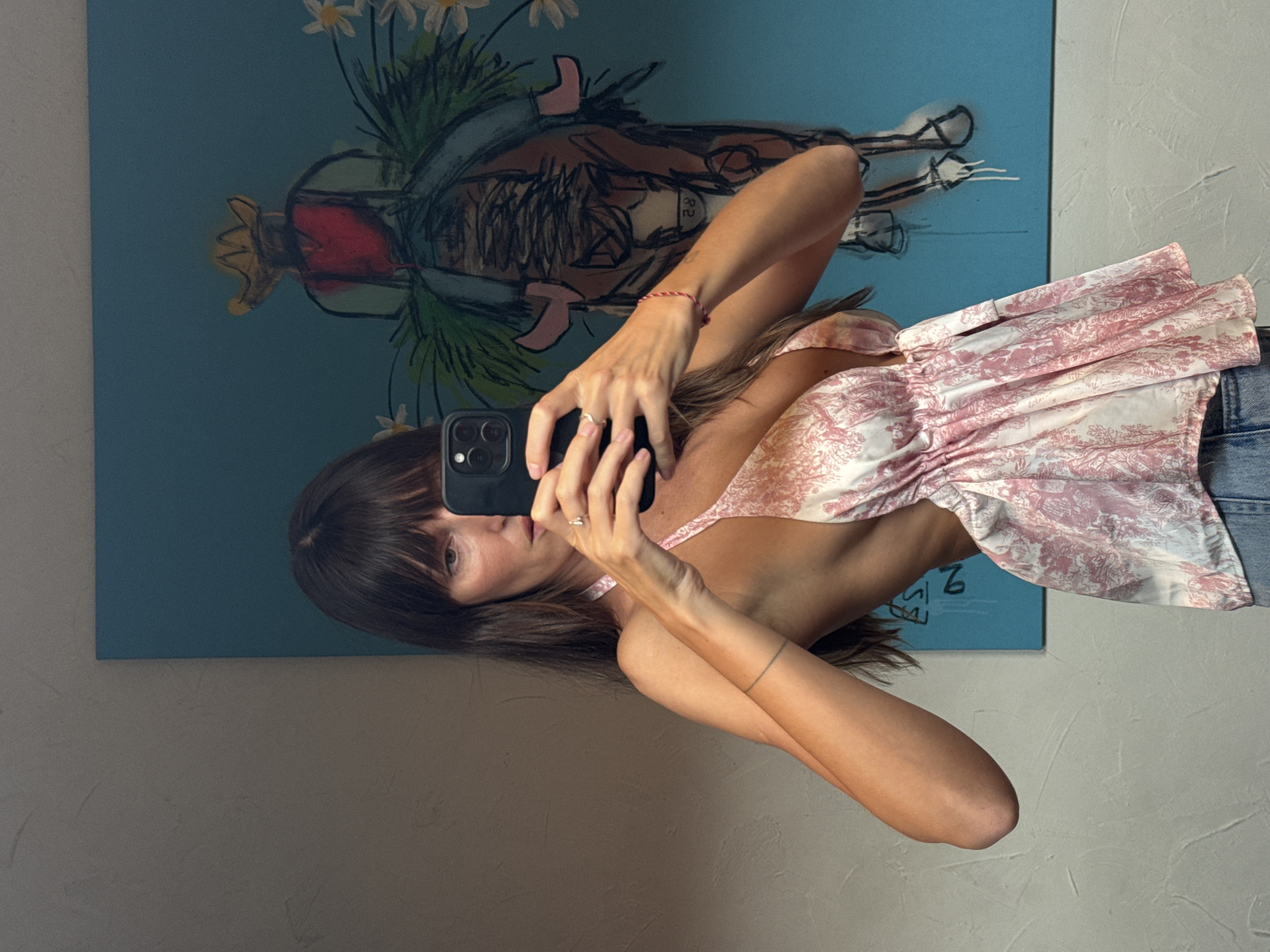 A woman is taking a selfie using a black smartphone in front of a blue painting. She is wearing a pink and white patterned top and blue jeans. The painting depicts a figure with flowers. The background consists of a light gray wall. The location appears to be indoors, with a casual and informal setting. The city is not identifiable from the image.