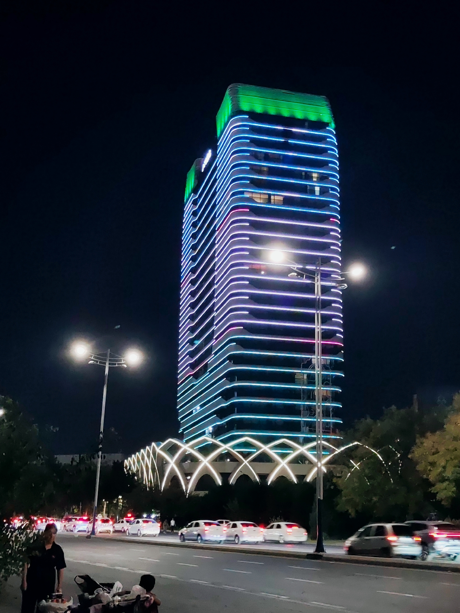 The image captures a nighttime scene in Tashkent, Uzbekistan, focusing on a tall, modern skyscraper illuminated with colorful LED lights that wrap around its exterior in horizontal bands of blue, purple, and pink. The building's top is highlighted with bright green lighting. In the foreground, a street is visible with several cars driving on it. To the left of the road, a woman stands near a cart laden with items, and a child sits on the ground behind the cart. Trees line the street on the left and right, and a large, arched light installation spans across the mid-ground, connecting to the building's base. Streetlights are also present, illuminating parts of the scene. The sky is dark, indicating it is nighttime.