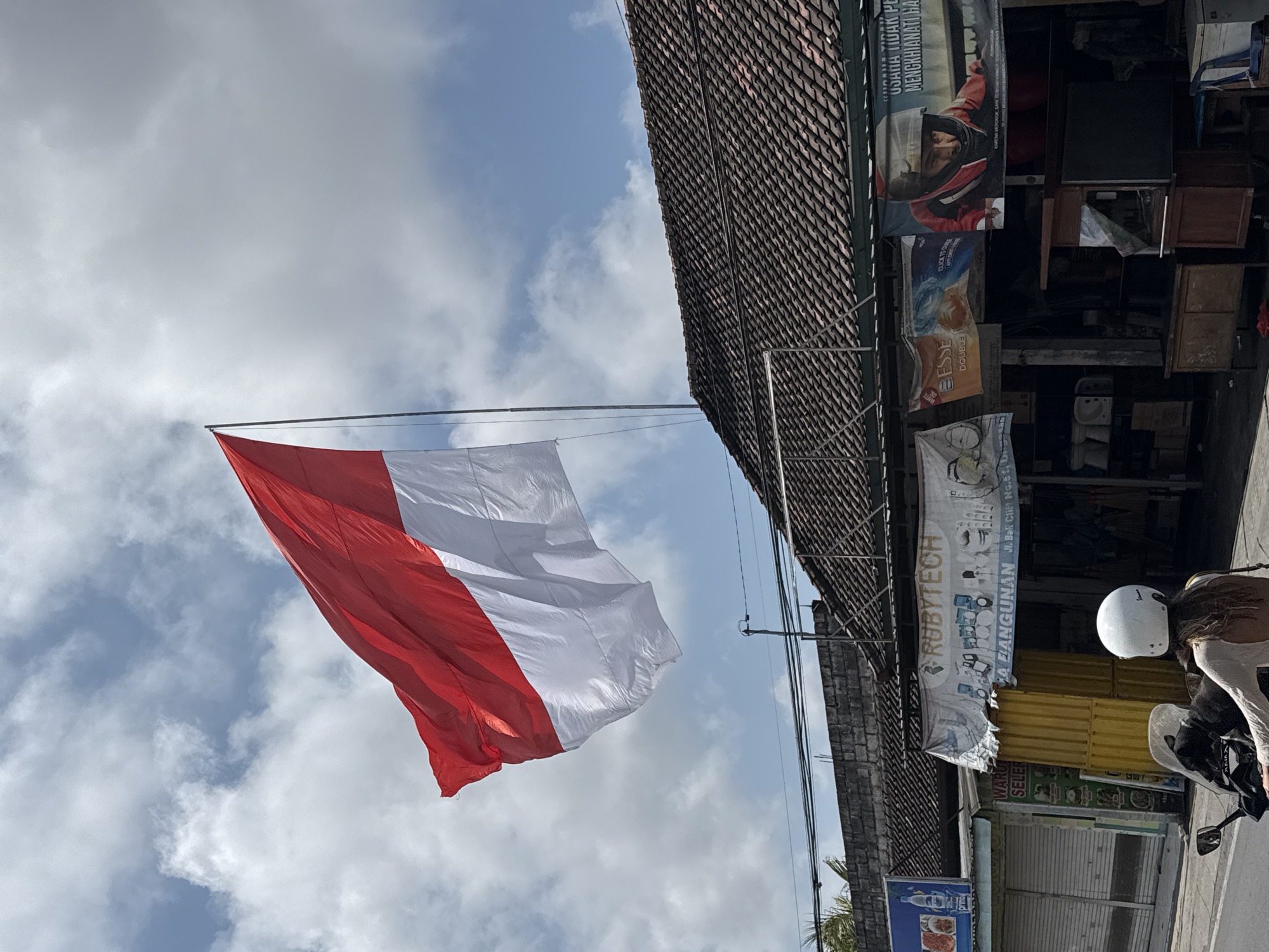In Kuta Selatan, Indonesia, a large Indonesian flag, red on top and white on the bottom, flutters against a cloudy sky from a flagpole mounted above a building. The building has a tiled roof and is lined with shops. Banners hang from the storefronts. One banner displays the name "RUBYTECH" along with images of various products. Another banner features a close-up of a person wearing a red racing helmet. In the foreground, a person on a white scooter wears a white helmet and a light-colored top, facing away from the camera. The street appears to be a commercial area with shops and stalls.