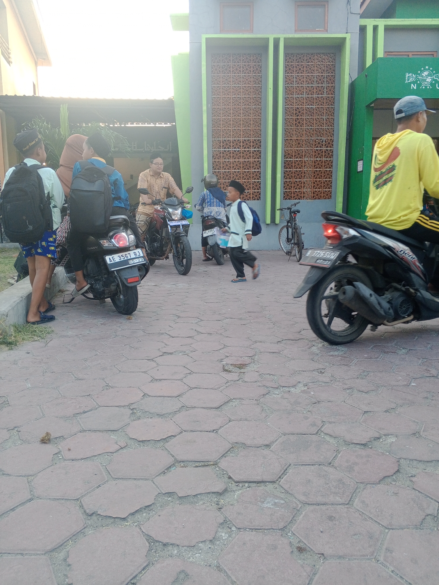 The image depicts an outdoor scene in Lembah, Indonesia, possibly near a building with green and brown architectural elements and decorative screens. The ground is paved with hexagonal red bricks. Several people are present, some on or near motorcycles.
On the left, three young males stand together, two of whom are wearing backpacks. One is wearing a black peci (a type of cap). Another person, a female wearing a hijab, stands behind them.
A man in a patterned shirt is seated on a motorcycle in the middle ground. Another motorcycle is visible to his right, ridden by a person in a blue patterned top. Further to the right, a young boy in a light blue shirt and dark pants, also wearing a peci, is walking away from the camera. He has a backpack on his shoulders.
In the foreground and mid-ground, a black motorcycle is prominent, with its rear wheel facing the camera. The license plate reads "AE 3567 BJ". Another motorcycle with a license plate "AE 4251 OG" is visible further back. A bicycle is parked to the left of the young boy. A person wearing a yellow hooded jacket is on a motorcycle to the far right of the frame, moving away from the camera. The overall lighting suggests it is daytime, with bright sunlight casting shadows.