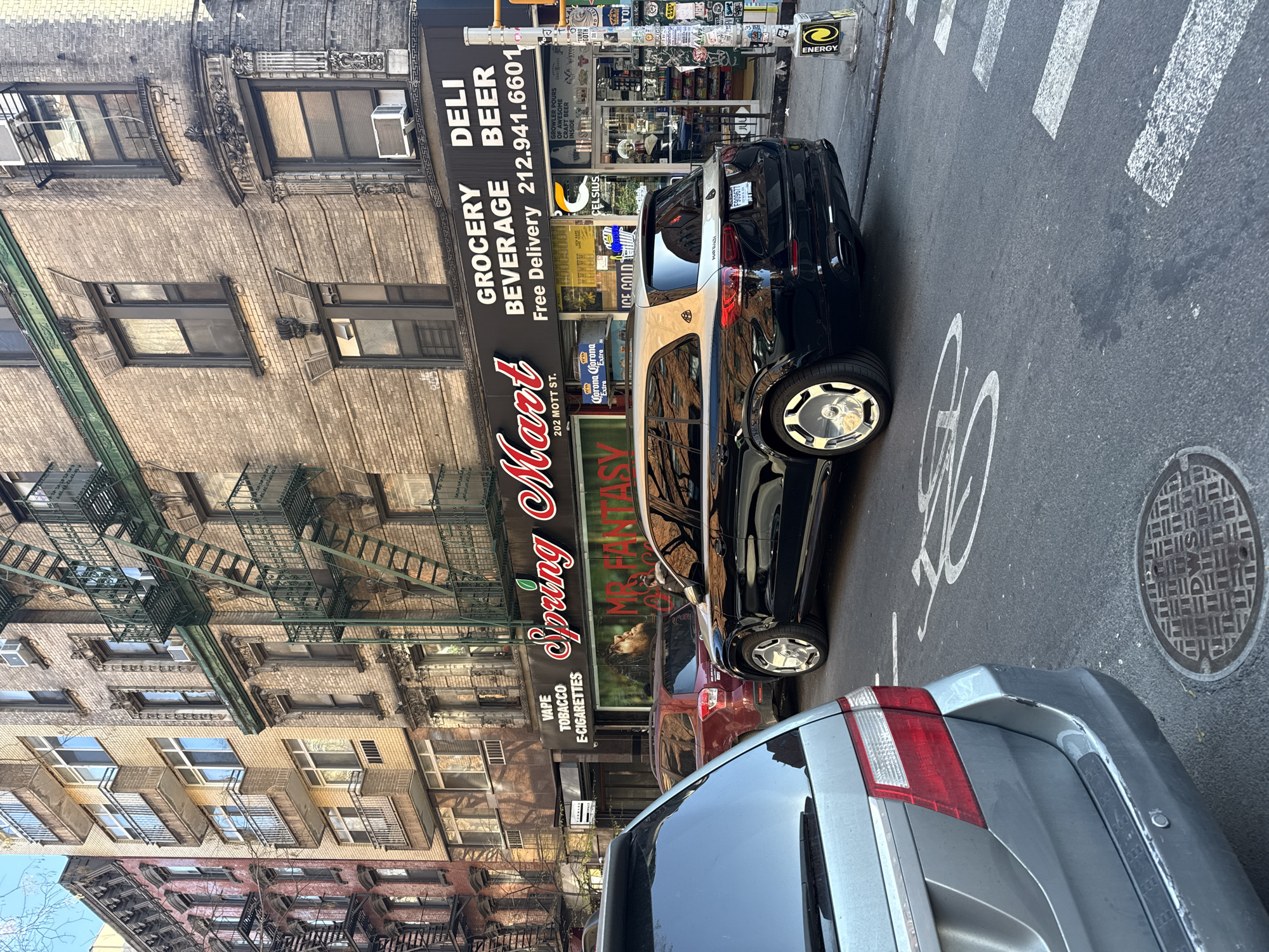 The image depicts a street scene in New York City. A black and white Maybach SUV is parked on the street in front of a storefront called "Spring Mart," which offers grocery, beverage, and deli items. Another storefront nearby offers vape, tobacco, and e-cigarettes. A silver car is parked behind the Maybach. The street is marked with a pedestrian crosswalk and a bicycle symbol. The buildings surrounding the scene have fire escapes and a typical NYC architectural style. The address "202 Mott St." is visible on the Spring Mart storefront, suggesting the location is on Mott Street.