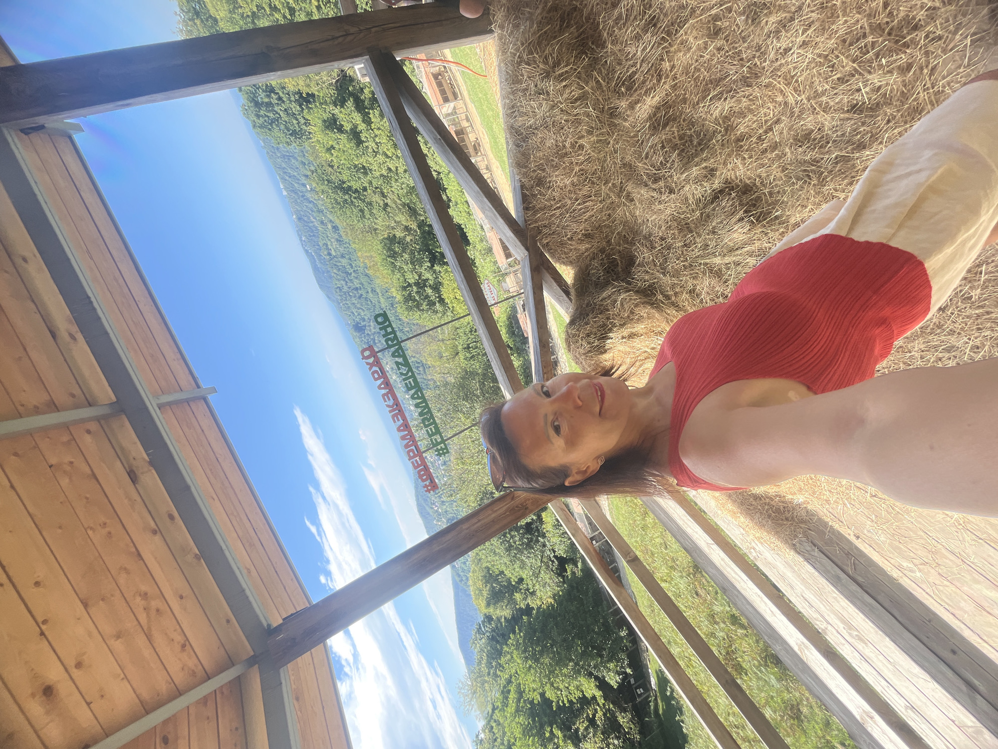 A woman in a red tank top and cream-colored skirt is taking a selfie in an outdoor structure with a wooden roof and railing. She is smiling at the camera. Behind her, a vast, green, mountainous landscape stretches to the horizon under a bright blue sky with scattered clouds. A large stack of hay bales is visible to her right. In the background, a sign with Cyrillic text and a hashtag can be seen. The location is identified as Progress, Russia.