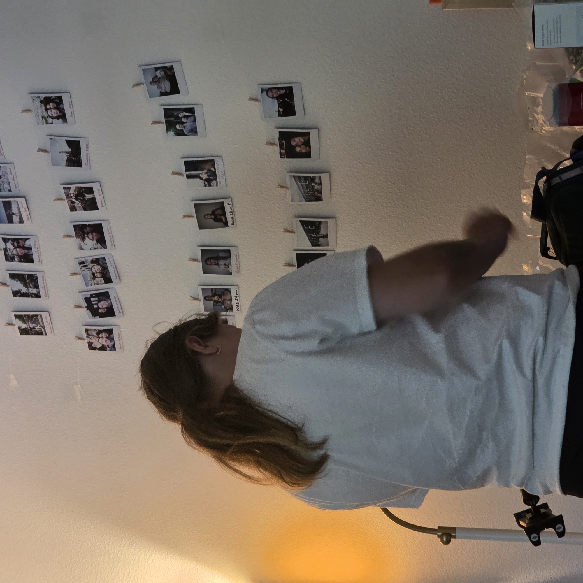 This media file shows a person in a room with a wall decorated with many small photographs clipped with clothespins. The photographs appear to be instant photos, possibly from a camera like a Polaroid or Instax. Some of the photos are of people, likely friends or family, posing together. Others show landscapes or abstract images. There are several rows of these photos, arranged in a grid-like pattern.
The person in the foreground is wearing a white t-shirt and has long brown hair. They are looking away from the camera, and their arm is raised, suggesting they might be interacting with something or someone out of frame, or perhaps adjusting a light or camera. The lighting in the room is warm and appears to be artificial, possibly from a lamp, casting a yellowish glow on the wall. This suggests it might be evening or nighttime.
On the right side of the frame, some objects are visible on a shelf or surface, including boxes and what appears to be a red container. A backpack is also partially visible. The wall has a textured surface. There are no obvious weather cues. The overall atmosphere seems casual and personal, reflecting a collection of memories displayed in a home environment. No visible text is legible in the photographs or elsewhere in the scene. The location context provided is Portland, United States of America, but there are no specific visual cues within the image that definitively confirm this location.
