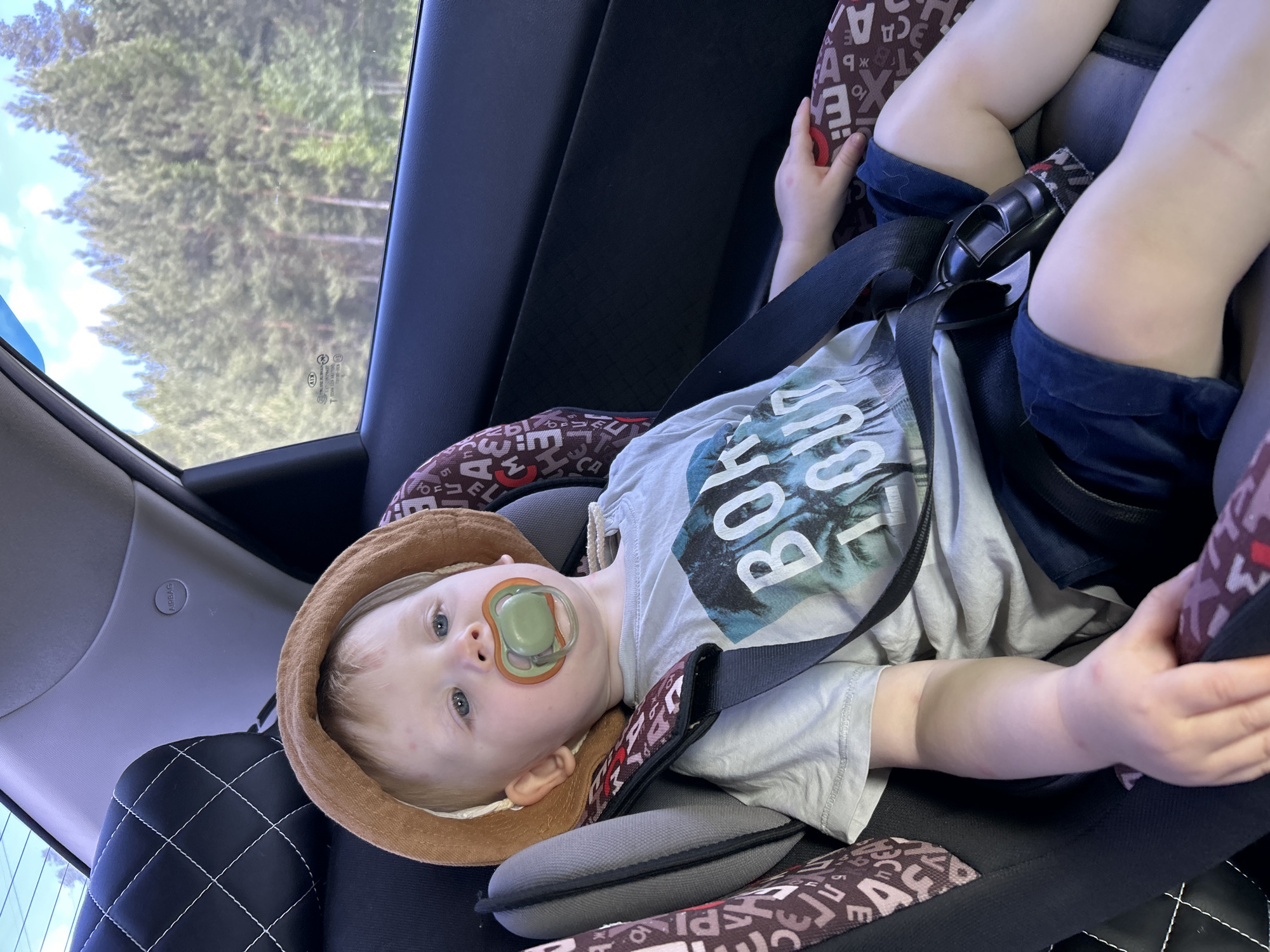 A toddler with fair skin, blue eyes, and blond hair is lying in a car seat. He has a green and orange pacifier in his mouth and is wearing a brown sun hat, a gray t-shirt with the word "BOLD" printed on it, and blue shorts. The car seat is positioned in the back of a vehicle. A window shows a view of green trees, possibly a forest. The exact location in Chaikovsky, Russia cannot be determined.