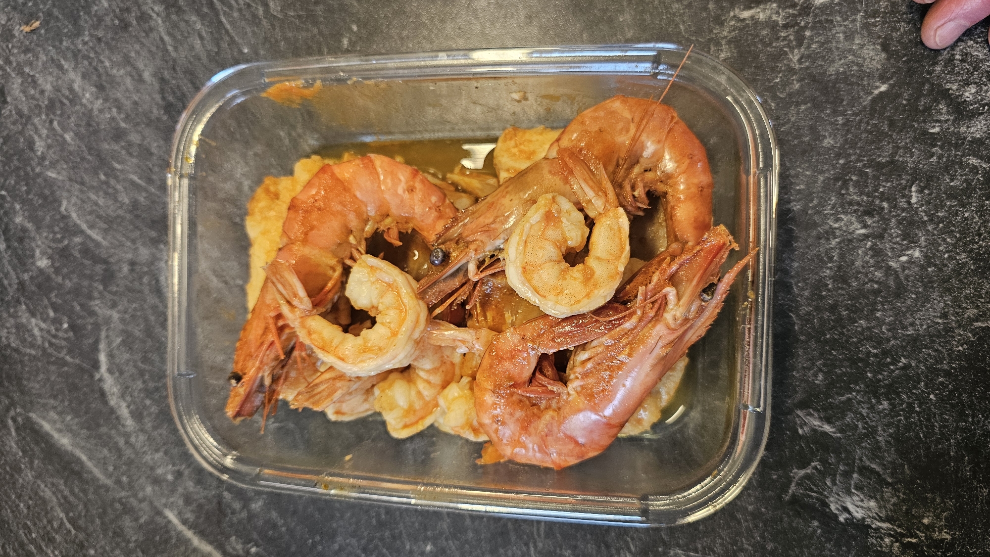 An overhead shot captures a clear, rectangular food container filled with cooked shrimp and a side dish, set against a dark, mottled background. The container, likely made of glass or plastic, holds numerous shrimp in various states of preparation. Several large, bright orange-red shrimp with their heads and shells intact are prominently displayed, characterized by their curled bodies and distinct antennae. Interspersed among these are smaller, shelled shrimp, revealing their lighter, opaque pink flesh.
A golden-brown sauce or liquid is visible at the bottom of the container, enveloping a yellowish-orange food item that forms the base of the meal. This base could be mashed potatoes, polenta, or a similar soft accompaniment. The container rests on a dark, textured surface, possibly a granite or marble countertop, which exhibits subtle grey and white striations. In the upper right corner, a small portion of a human hand, including fingertips and a thumb, is visible, suggesting interaction with the container, perhaps holding or placing it. The image depicts a prepared meal, ready for consumption or storage, with focus on the vibrant colors and fresh appearance of the seafood. No text is visible in the image. The setting appears to be an indoor environment, such as a kitchen or dining area.