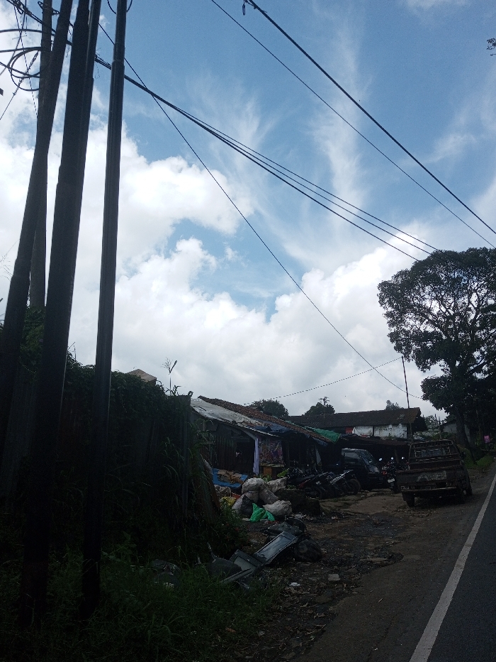 The image depicts a roadside scene in Ura-Dozhi, Bhutan, under a blue sky with scattered white clouds. On the left, three dark, vertical utility poles rise from the foreground, supporting multiple black electrical wires that extend diagonally across the upper frame. Below the poles, a dense mass of dark green foliage covers an unseen structure or embankment. To the right of the foliage, a collection of informal structures with corrugated metal and tarp roofs lines the roadside. In front of these structures, piles of white and green bags, along with discarded items including what appears to be a broken television screen, are visible on the uneven ground. A dark pickup truck and several motorcycles are parked off the paved road, which features a white edge line and runs diagonally from the bottom right. In the background, traditional white-walled buildings with dark roofs are partially obscured by a large, leafy tree on the right side of the frame. No individuals are visible in the scene.