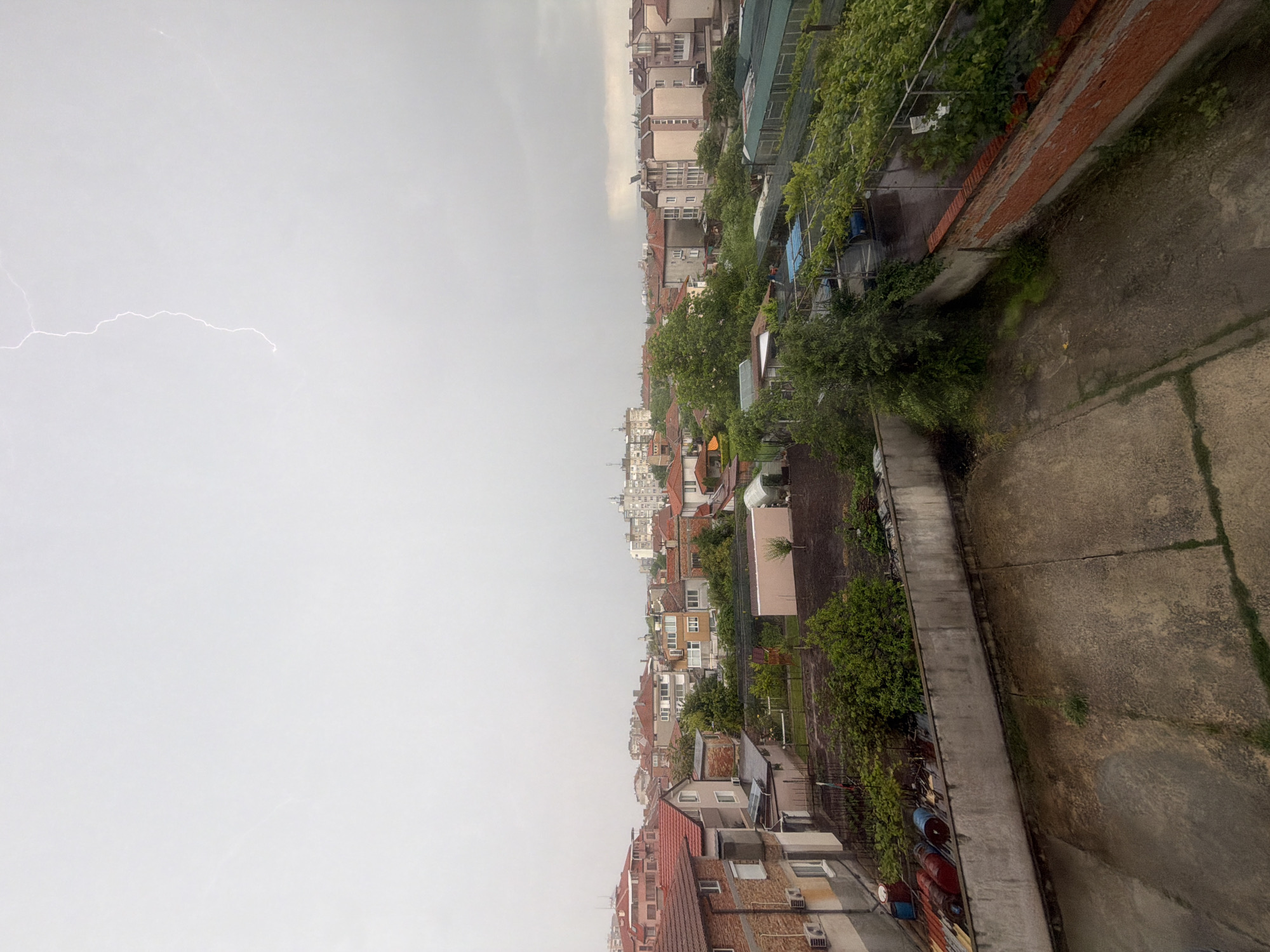A view from an elevated position looking out at a city, likely Burgas, Bulgaria, based on the buildings and the prevalent overcast sky. The houses have brick and orange tile roofs, with some modern buildings interspersed with older structures. Green foliage is scattered throughout, suggesting gardens or yards. On the left, lightning flashes across a uniform gray sky, indicating a storm. The right edge of the image shows a concrete wall or structure, possibly a balcony or rooftop edge, with additional greenery.