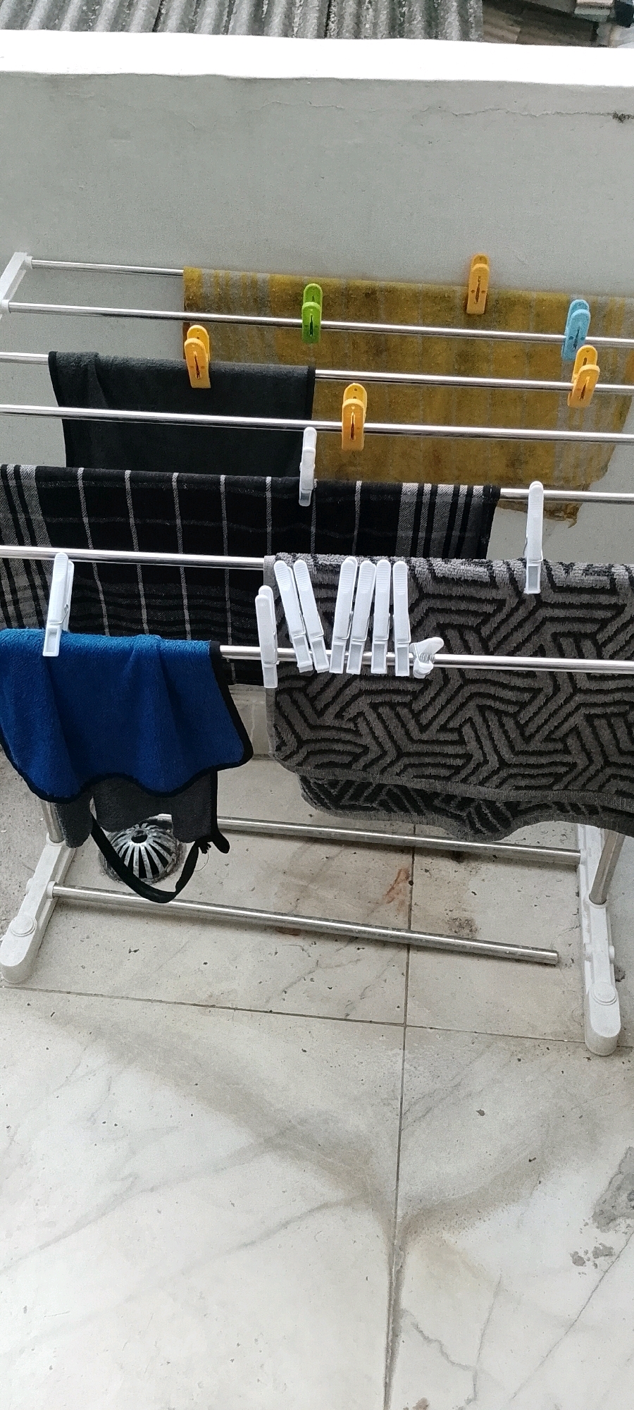 An outdoor clothes drying rack is shown with several items of clothing hanging on it. From top to bottom, there are two yellow towels, a dark-colored towel, and a patterned towel. A blue towel is also hanging on the left side. The drying rack is positioned on a tiled surface, possibly a balcony or patio, with a white wall and a corrugated roof visible in the background. Various colored clothespins are used to secure the clothing. The scene is lit by natural daylight. There are no people visible in the image.