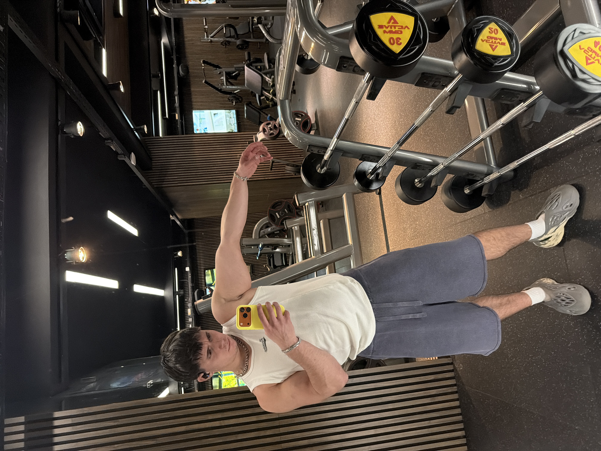 A man is lying on his back on a striped surface in what appears to be a gym in Sofia, Bulgaria. He is wearing grey athletic shoes, white socks, blue shorts, a white sleeveless shirt, and has a chain around his neck. He is holding a yellow smartphone, seemingly taking a selfie. His left arm is raised, and he has a silver bracelet on his left wrist. The gym equipment, including weight racks with labeled weights, is visible in the background. The lighting is bright and artificial. There are television monitors and exercise machines visible as well.
