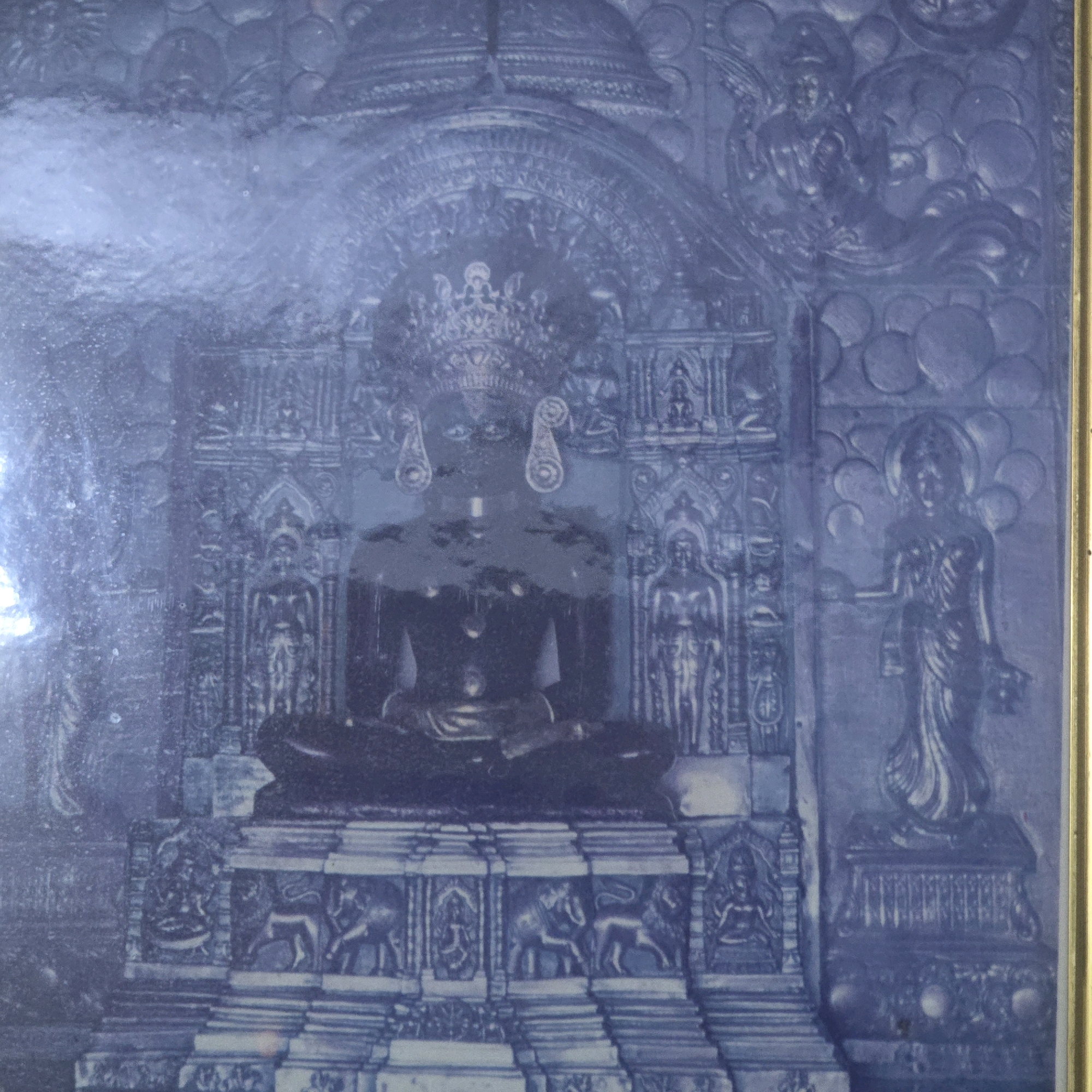 The image depicts a detailed bas-relief sculpture in a blue monochrome tone, framed by a gold border. The central and dominant subject is a seated figure, likely a deity or revered personage, adorned with an elaborate headdress and large earrings. This figure is positioned within an ornate architectural structure, possibly a shrine or temple facade, characterized by intricate carvings and decorative motifs. Flanking the central figure are smaller, standing figures, also depicted in relief. The lower portion of the sculpture features a tiered base with repeating patterns, including what appear to be depictions of elephants and possibly lions. To the right of the central figure, another standing figure is visible, in a graceful pose. The overall style suggests a religious or mythological theme, with the detailed carvings indicating significant craftsmanship. The location is identified as a Jain temple in Palghar, India. The artwork appears to be a photographic reproduction of a carved relief, possibly made of stone or metal, given the monochromatic blue hue.