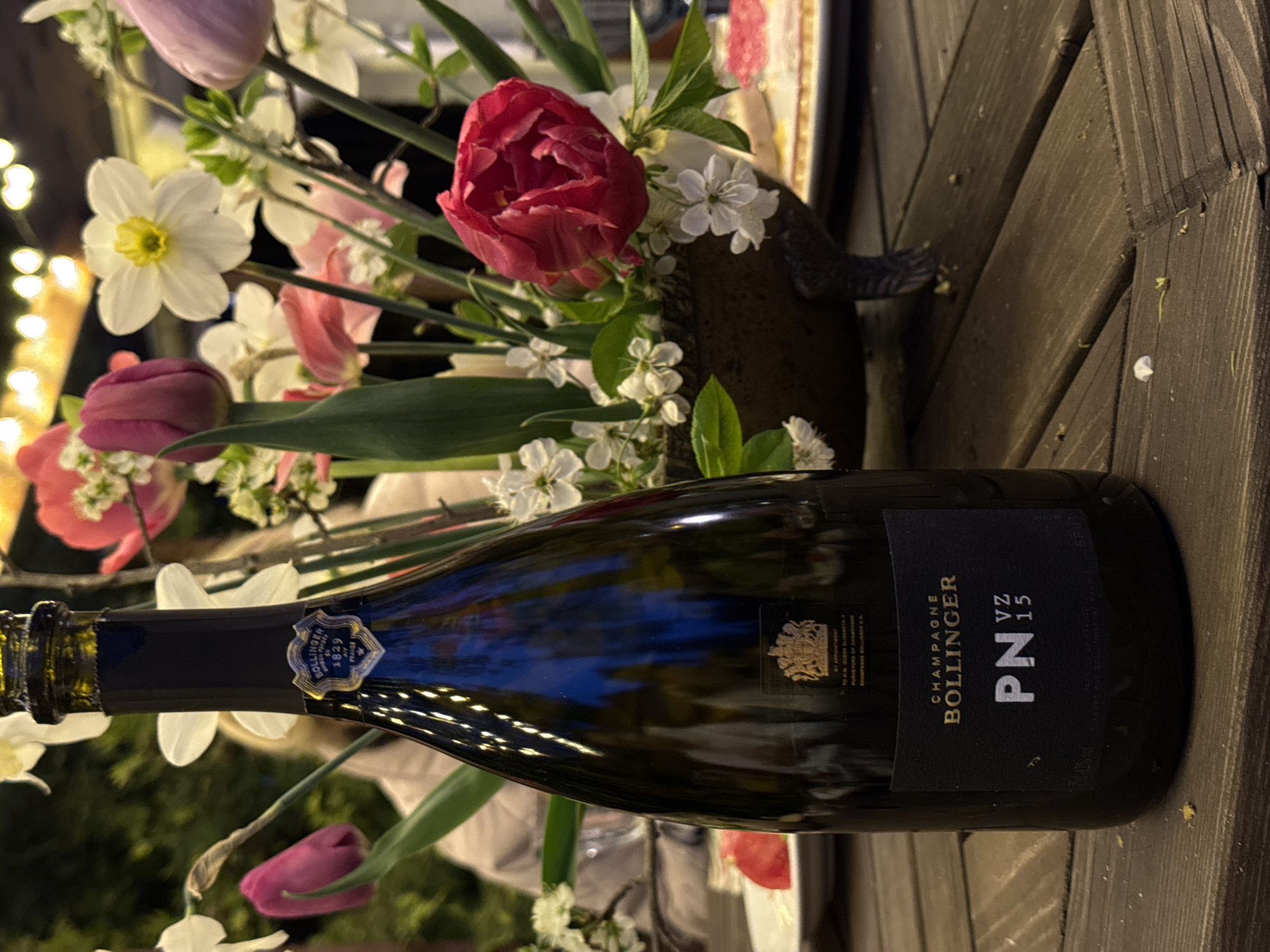 A medium shot depicts a bottle of Bollinger champagne on a dark, wooden, planked surface with a flower arrangement behind it. The champagne bottle, labeled with "CHAMPAGNE BOLLINGER PN VZ 15", is oriented horizontally, with its label facing the viewer. Behind the bottle is a vibrant flower arrangement with tulips, daffodils, and other smaller white flowers in a dark-colored vase. In the background, bokeh lights are visible, suggesting an outdoor setting. The overall atmosphere suggests a celebratory event. The city of Sapronovo, Russia, is not directly discernible from the image.