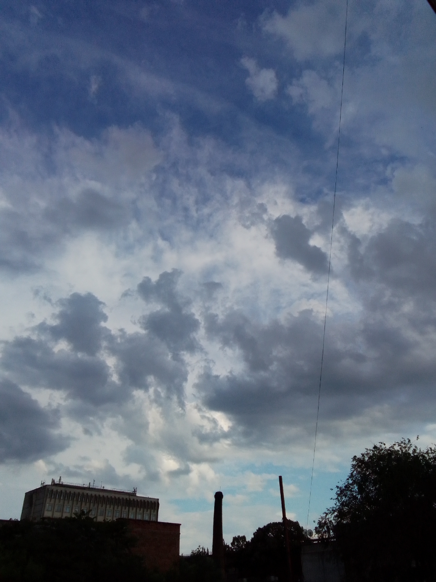 This is a medium-angle, outdoor, low-light, vertical shot of a cloudy sky above an industrial area. The sky is filled with various shades of gray and white clouds, with patches of blue sky peeking through. A thin black wire runs vertically through the center of the image. In the foreground, silhouetted against the sky, are parts of buildings and trees. On the left, a large, multi-story building with a flat roof and rows of windows is visible. To the right of it, a brick building and a tall, slender smokestack can be seen. In the far right, the dark silhouette of dense foliage from trees obscures a portion of the sky. The overall atmosphere is somber due to the overcast sky and the industrial setting.