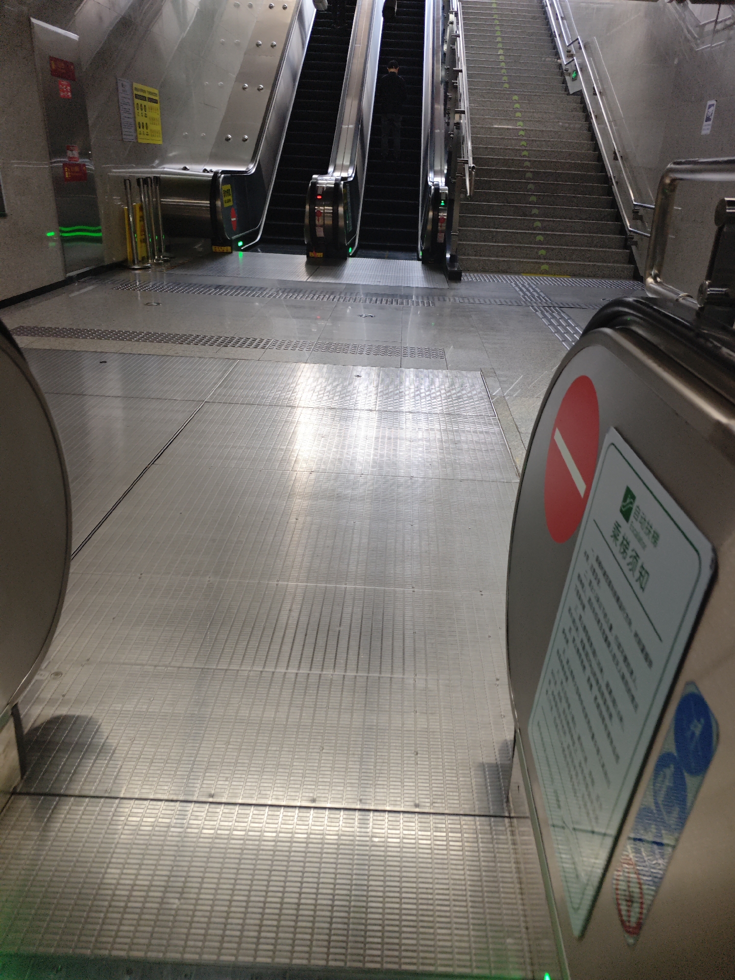 The image captures a view from the top of an escalator in what appears to be a transportation hub, most likely a subway station, in Liangjiang, China. The scene includes two sets of escalators, one descending and another adjacent to a traditional staircase. A person can be seen on the descending escalator. Noticeable are the safety and information signs in Chinese. The environment is brightly lit, giving a clean, modern impression of the location. The dominant material appears to be stainless steel or aluminum, widely used for the escalators and floor panels.