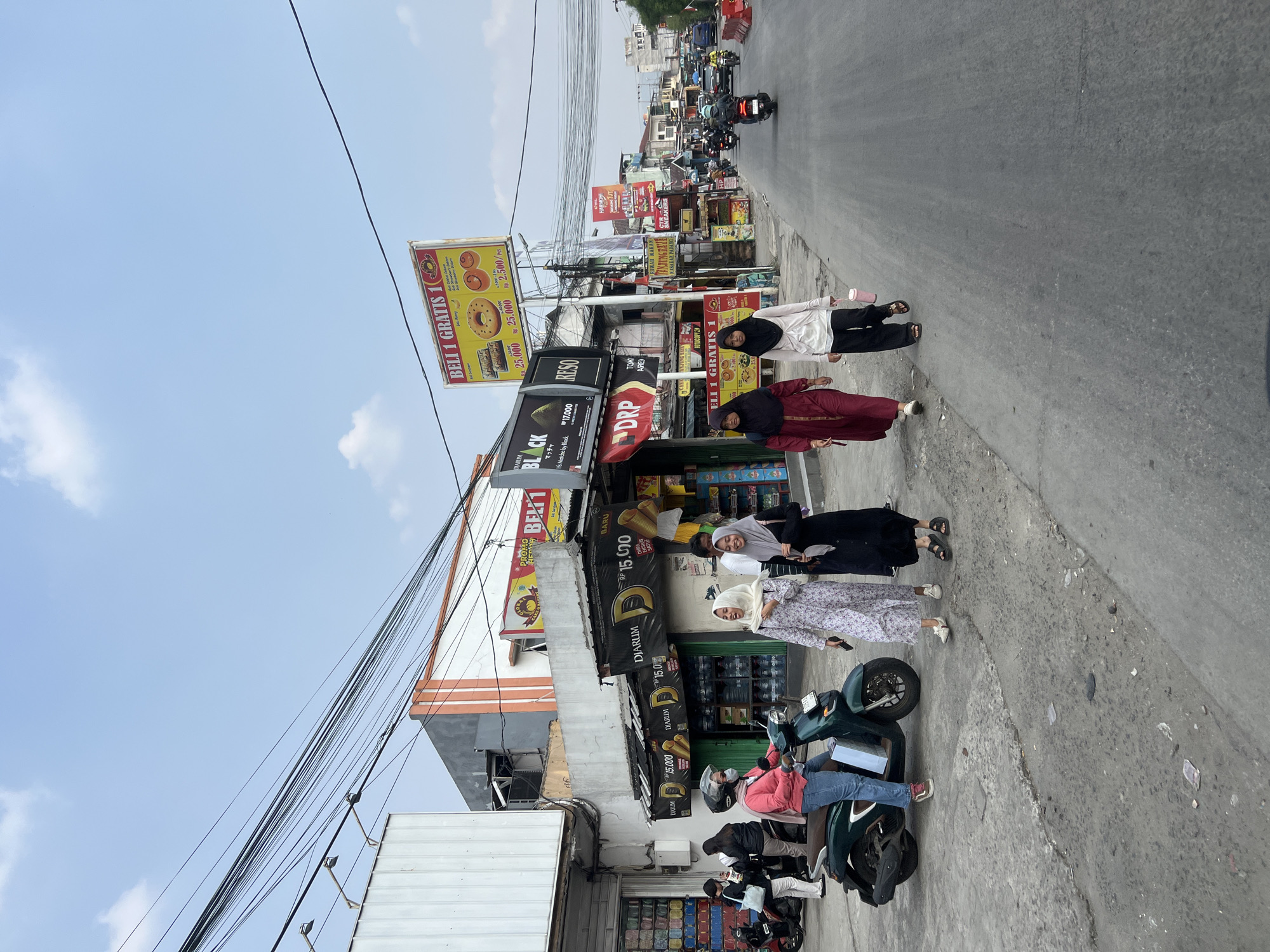 A street-level view captures a section of a bustling street in Citeureup, Indonesia, under a partly cloudy sky. The scene is dominated by a row of buildings lining the left side of the road, characterized by their varied architectural styles and commercial signage. Prominently displayed are advertisements for "Djarum Black" cigarettes, "Reso," and a "Beli 1 Gratis 1" (Buy 1 Get 1 Free) promotion for donuts and pastries, indicating a retail and food-oriented area. Electrical wires crisscross overhead, a common sight in urban Indonesian environments.
On the sidewalk, several individuals are present. Four women are walking along the street. One, wearing a pink jacket and blue jeans, is seated on a green scooter. Another woman in a floral dress and hijab is walking, as are two other women, one in a maroon abaya and hijab, and another in a white hijab and dark pants. To the left, a group of people are gathered near the entrance of a store, some holding bags. A motorbike is parked further down the sidewalk.
The street itself is paved with asphalt and shows signs of wear and tear, with some debris scattered on the surface. Motorbikes and a few cars are visible further down the road, suggesting ongoing traffic. The overall impression is of a typical urban street scene in a developing area of Indonesia, with commercial activity and pedestrian and vehicular traffic.