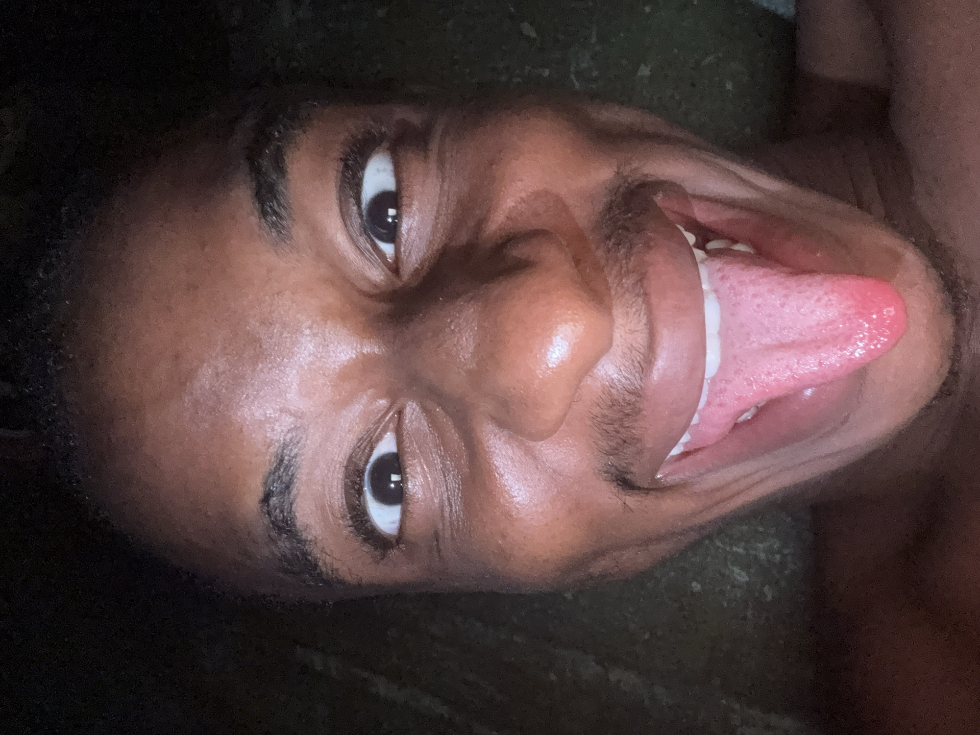 The image is a close-up, low-angle shot of a young Black male with dark skin, sticking out his tongue. The lighting is dim and appears to be artificial, casting shadows on his face. His eyes are wide open, and he has a playful expression. His tongue is extended and appears to be pink. The background is dark and out of focus, with some texture suggesting it might be a rough surface like pavement or dirt. There are no discernible landmarks or elements that would indicate a specific location within Nkpor, Nigeria.