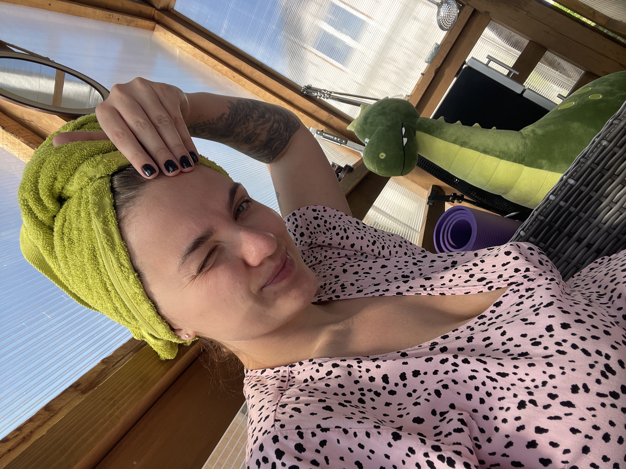 A woman is indoors, possibly in a greenhouse, wearing a pink and black patterned top and a green towel wrapped around her head. She is winking with her left eye and has her right hand on her forehead. She has dark nail polish and a tattoo on her right arm. Next to her is a large green alligator plush toy and a purple yoga mat. Behind the alligator, various objects are partially visible including a skimmer, and part of a window with blue sky visible through it. The structure is made of wood and transparent material. The location is Мосметаллоконструкция Онт, Russia.