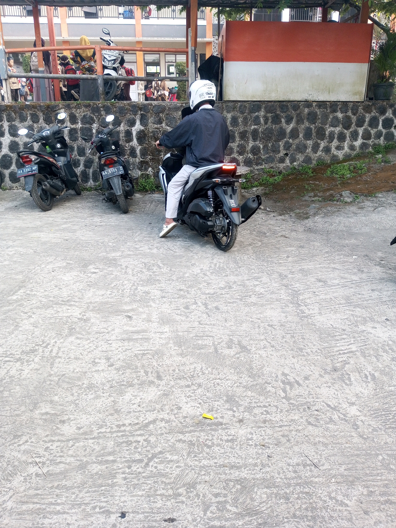 In Krasak, Indonesia, a person wearing a white helmet and a dark long-sleeved shirt and white pants is seated on a dark-colored scooter. The scooter has its brake light illuminated. The scooter is parked in front of a stone wall. Behind the wall, there is a building with a red and white horizontal sign. In the foreground, other dark-colored scooters are parked on a concrete surface. The environment appears to be outdoors, possibly a parking area or school campus.