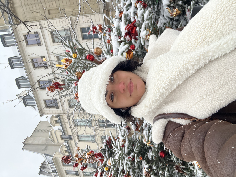 An image taken outdoors depicts a woman in a snowy Parisian setting. The woman is centered, looking directly at the camera with a slight smile, suggesting a selfie. She wears a white, fluffy bucket hat, a matching white, fluffy scarf, and a brown shearling-lined jacket. Dark, curly hair is visible around her face. Her cheeks show a slight flush.
Behind her, several snow-covered evergreen trees are visible, decorated with red bows and gold and red spherical ornaments, indicating a festive, possibly Christmas, display. Snow lightly covers the branches of deciduous trees. In the background, classic multi-story Parisian buildings with light-colored facades and grey mansard roofs are prominent. The rooftops are covered with snow, and dormant windows are visible. The sky is overcast and uniformly light grey, consistent with snowy conditions. The scene captures a winter day in a public area of Paris, France.