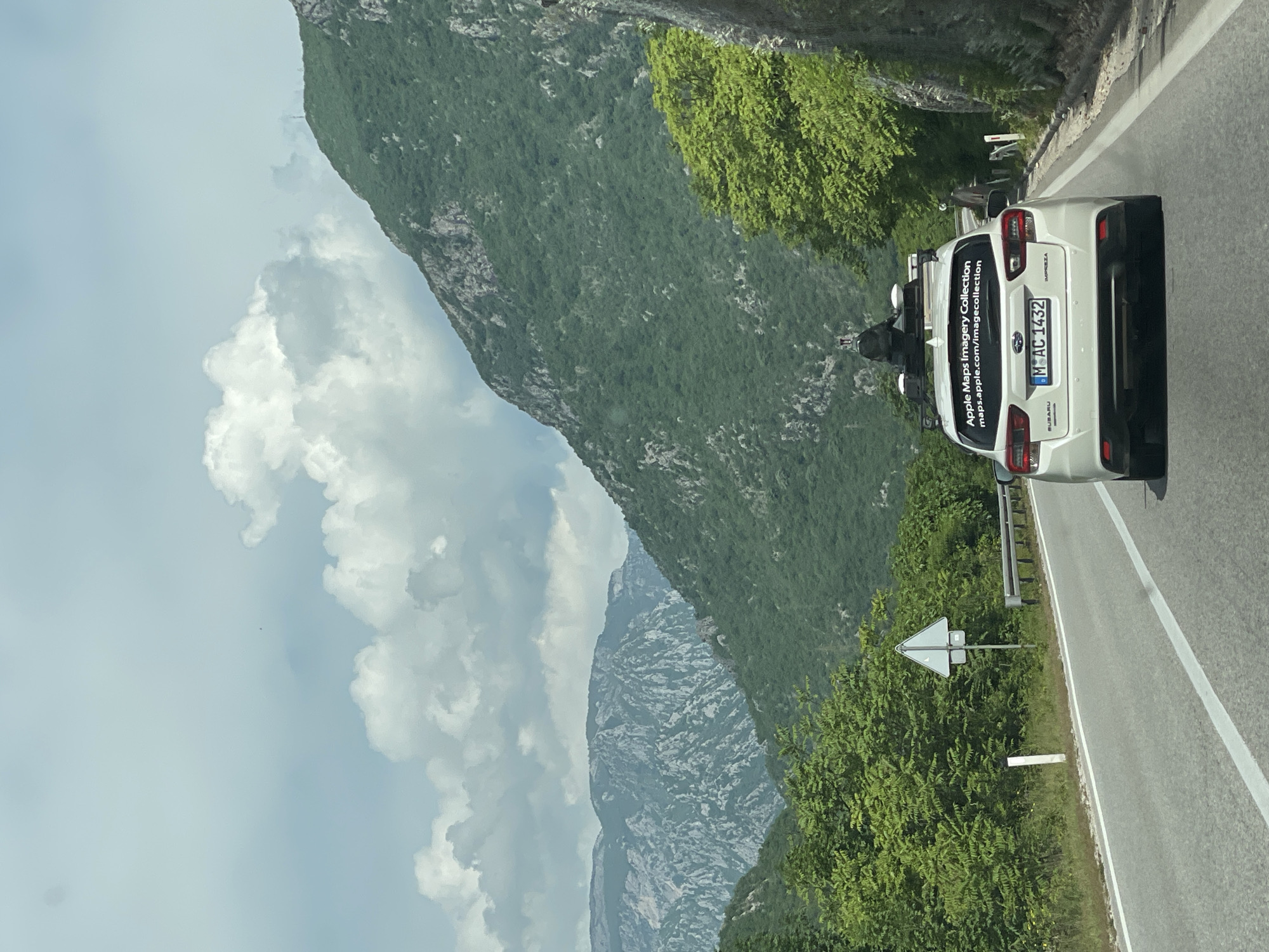 A white Subaru Impreza with the license plate "MO AC 1432" is parked on the right side of a paved road. The car features an "Apple Maps Imagery Collection" sticker on the back. The road runs alongside a lush, green mountain. The sky is visible with scattered clouds. The location is in the vicinity of Donji Jasenjani, Bosnia and Herzegovina, based on the probable location of Apple Maps imagery collection. The view is seen from another vehicle, indicating the photo was taken by someone traveling on the road.