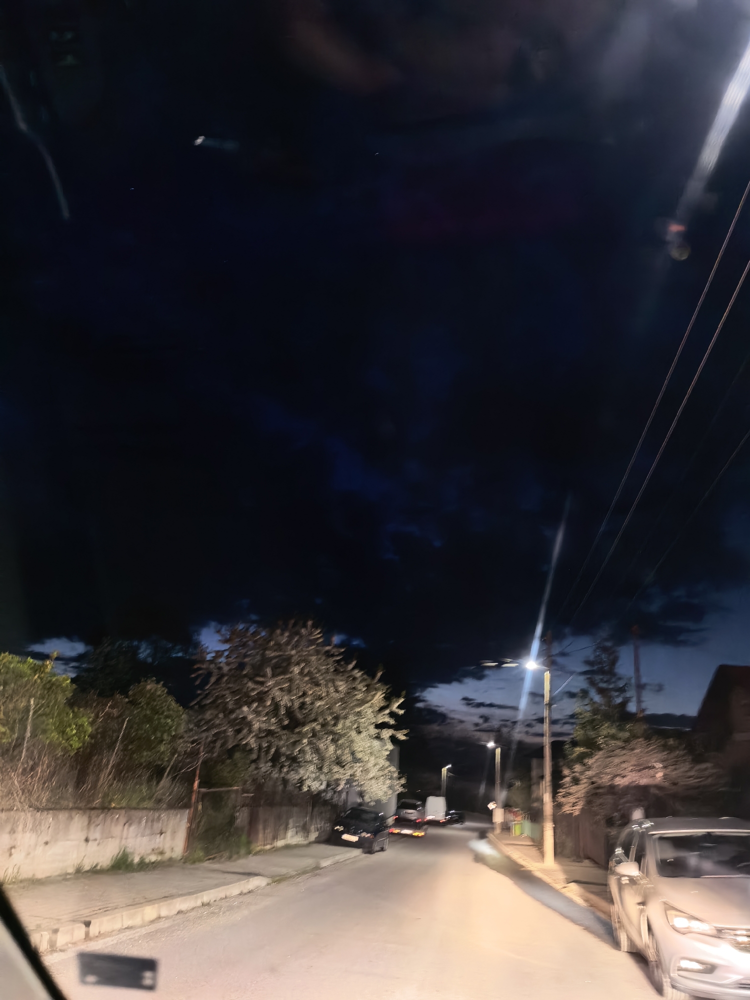 The image displays a nocturnal or late dusk scene of a street, viewed from inside a vehicle, likely through a windshield given the reflections. The sky is predominantly dark, almost black, filled with heavy clouds, though lighter blue-grey areas are visible through breaks in the clouds, particularly towards the horizon.
A paved road, possibly wet or reflective, curves slightly as it extends into the background. Streetlights are illuminated, casting bright, artificial light onto the road and the immediate surroundings. On the right side of the foreground, a light-colored car, possibly silver, is parked with its front visible. Further down the street, a dark car is parked on the left, and a white van and another vehicle are discernible in the distance.
Both sides of the road are bordered by dense vegetation, including trees with lighter-colored foliage and bushes. A low concrete wall and a fence run along the left side of the street. Prominent utility poles with overhead wires stand on the right, silhouetted against the dark sky. A bright streetlight on the right side casts a strong vertical beam of light, while another streetlight illuminates the street further down on the left.
The scene depicts a quiet, residential or semi-rural setting after dark. There are no people visible. No discernible text can be found in the image.