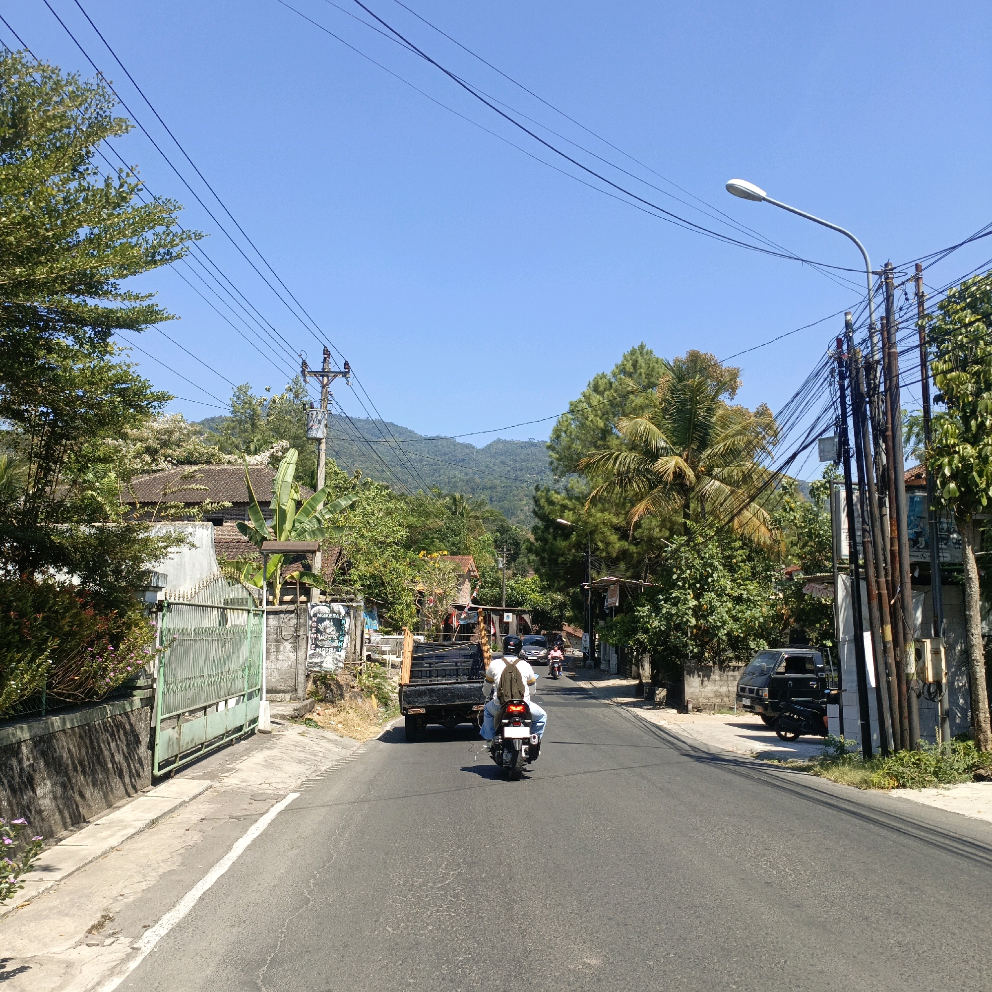 The image depicts a street scene in Krajansari, Indonesia, on a clear, sunny day with a bright blue sky. The perspective is from a moving vehicle on a paved road that curves gently to the right. On the left side of the road, a green metal gate is open, revealing a stone wall and a building with a tiled roof, surrounded by lush green foliage and trees, including a large banana tree. A smaller building with a red-tiled roof is visible further down the street. On the right side of the road, a black pickup truck is parked on the side, with a motorcycle parked in front of it. A utility pole with numerous black wires is prominent on this side, along with a street light.
The road is two lanes wide and shows some wear and tear. Several vehicles are present: a black pickup truck in the foreground left, a black pickup truck parked on the right, and two motorcycles. The first motorcycle, in the center of the road, is ridden by a person wearing a white shirt and blue jeans, with a backpack. Another motorcycle is further down the road, carrying a single rider. In the distance, rolling green hills and mountains are visible under the clear sky. The environment appears to be a semi-rural or suburban area with a mix of residential buildings and natural vegetation.