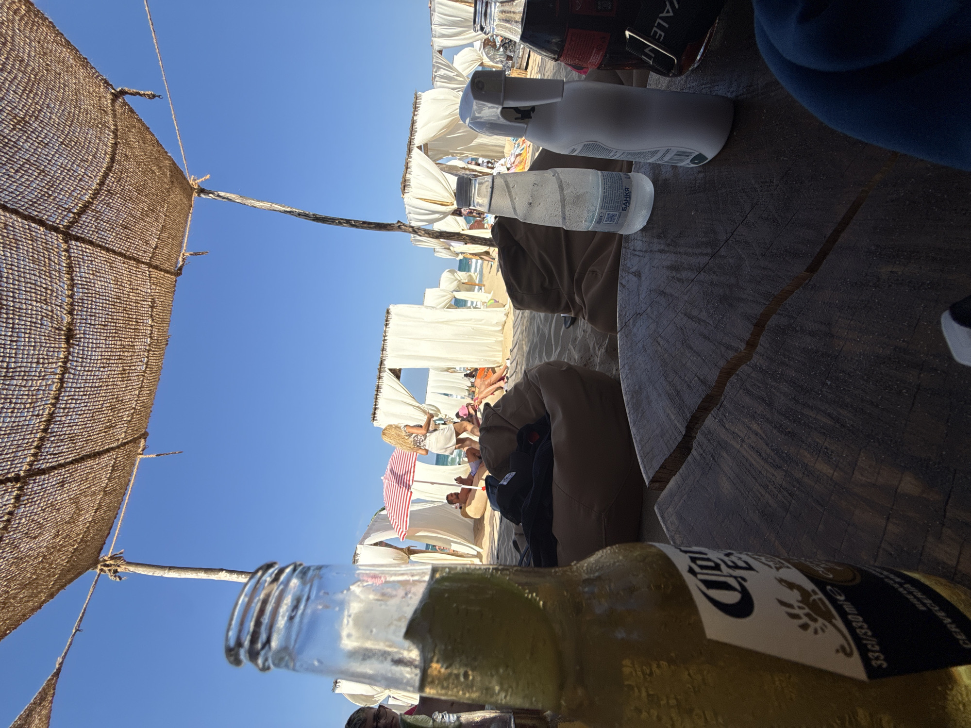 An eye-level, medium shot captures a sunlit beach scene at the North Beach in Primorsko, Bulgaria. In the foreground, a wooden table is set with a condensation-covered bottle of Corona Extra beer, a clear bottle of water, and a white spray bottle. Behind the table, several white cabanas with open fronts line the sandy beach, each draped with sheer white curtains. A red and white striped umbrella is set up between two cabanas, and a few people are visible relaxing on beanbag chairs and sitting in the sand. In the background, the clear blue sky meets the horizon of the Black Sea. The overall atmosphere is one of leisure and relaxation.