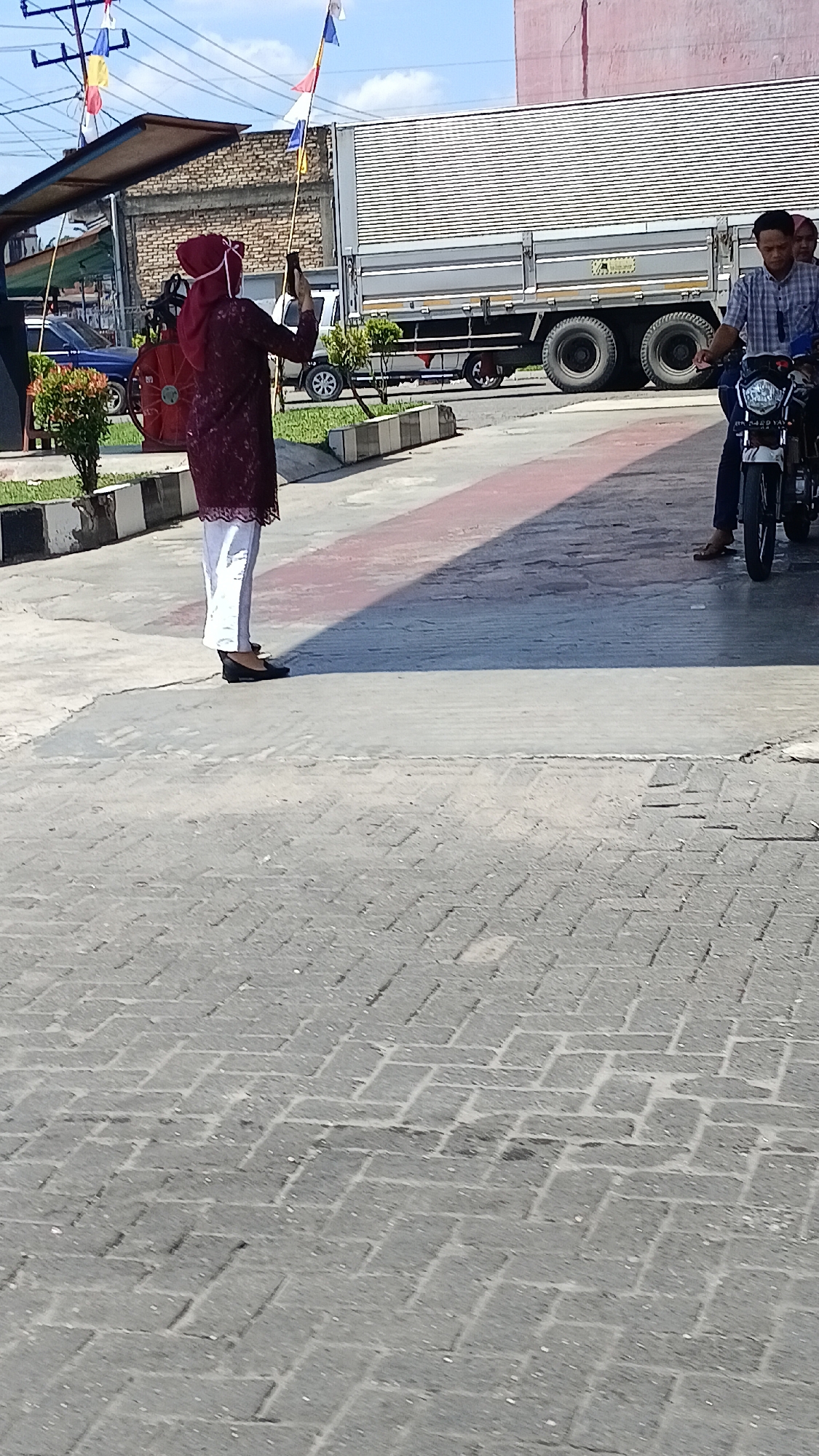 In Aek Ledong, Indonesia, a woman wearing a burgundy hijab and a patterned maroon top with white pants stands on a paved surface, holding a smartphone in her right hand and a flag pole with small colorful flags in her left. She is facing away from the camera, appearing to take a photo or video. To her left, a blue car is parked. Behind her, a large silver truck with a white top is parked. To the right of the truck, two men are on a motorcycle, with the driver wearing a checkered shirt. The environment includes a building with a pink facade and a blue sky overhead with some clouds. The ground is paved with interlocking bricks and concrete.