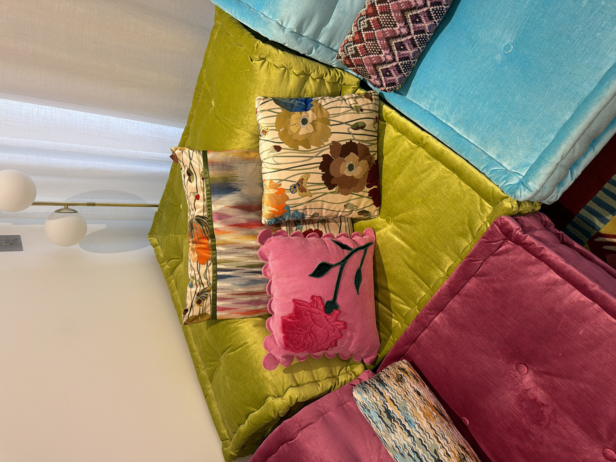 The image displays a collection of decorative pillows and cushions arranged on a surface, possibly a bed or seating area. The color palette is vibrant, featuring hues of green, pink, blue, and brown. Various textures and patterns are evident, including floral motifs, geometric designs, and textured fabrics. The pillows are placed in a seemingly random but visually appealing arrangement, adding a touch of eclecticism to the setting. A lamp with two white globes is visible in the background, suggesting an indoor environment. The scene doesn't definitively place it in London or anywhere in the United Kingdom.