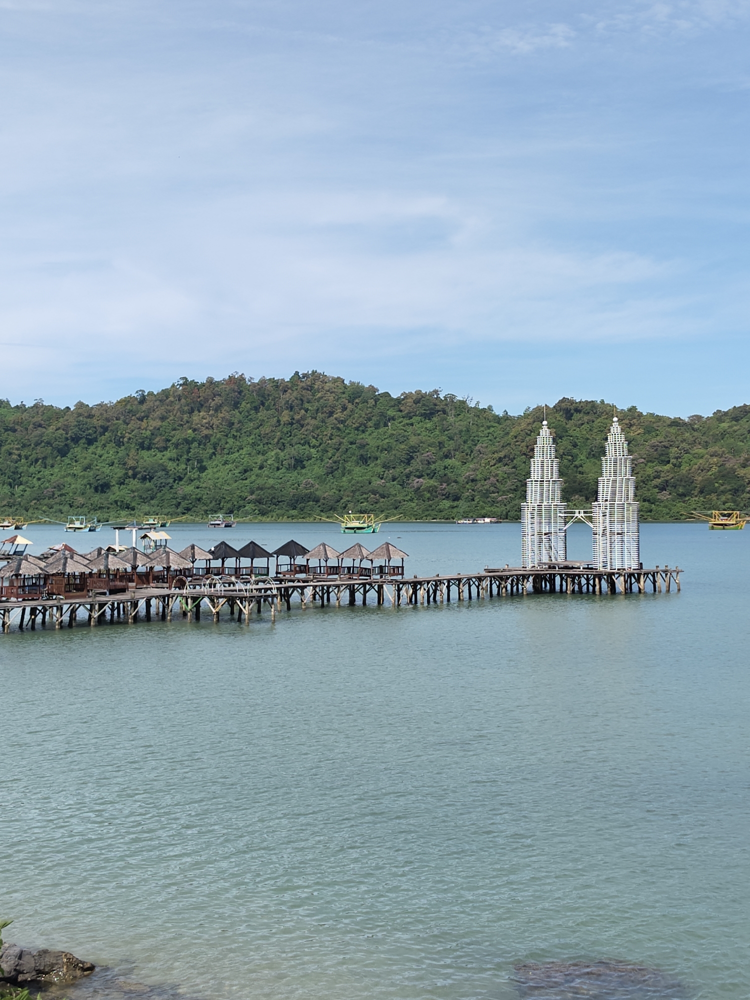 The image depicts a waterfront scene in Layeun, Indonesia, featuring a wooden pier extending into the ocean. The pier has small, thatched-roof pavilions along one section and culminates in a structure resembling twin towers, constructed from white, open-framework material. In the background, a forested hillside provides a backdrop to the calm, light-blue water. Scattered boats are visible on the water's surface. The sky is partly cloudy. In the lower foreground, rocks sit at the edge of the shoreline.