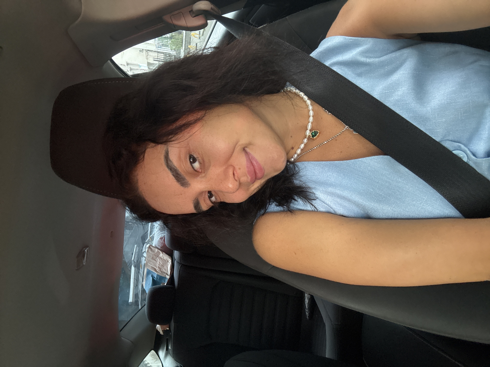 A young woman with dark, wavy hair and brown eyes is seated in the passenger seat of a car, looking towards the camera with a slight smile. She is wearing a light blue sleeveless top and a pearl necklace with a green pendant. A black seatbelt is fastened across her chest. The interior of the car is visible, including black leather seats and the headrest. Outside the car window, buildings and vegetation can be seen, suggesting an urban environment. The background suggests the scene is in the city of Tbilisi, Georgia.