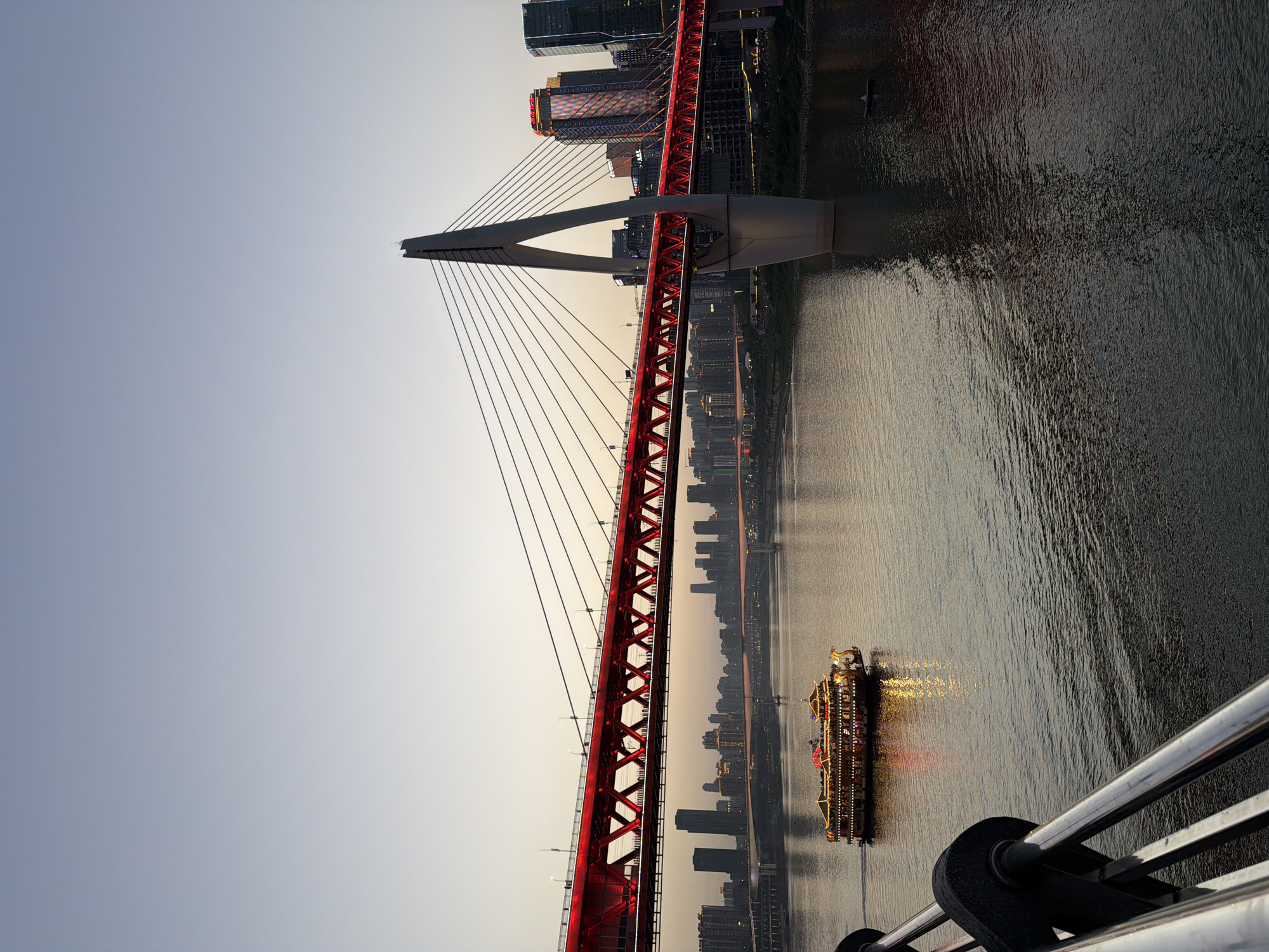The vertical image captures an urban river scene during twilight in Yuzhong District, China, likely in Chongqing. A prominent cable-stayed bridge spans diagonally from the lower left to the upper right, its main truss painted a striking red and appearing to be illuminated. A grey pylon rises towards the upper center, from which numerous cables descend to support the bridge deck.
On the left side of the frame, a clear, pale blue-grey sky dominates, subtly transitioning to a brighter hue towards the right. On the right side, a wide river reflects the soft light from the sky and the emerging artificial illumination from the city. A brightly lit passenger boat, adorned with numerous warm yellow and some red lights, navigates the river, casting shimmering reflections on the water's surface.
In the background, along both sides of the river, a dense cityscape of high-rise buildings and skyscrapers is visible. Many of these structures display illuminated windows and facade lighting, signifying the transition from day to night. A partial metal railing is visible in the bottom right corner, suggesting the photograph was taken from an elevated viewpoint, such as another bridge or a high building. The scene conveys the grandeur and activity of a vibrant city during the atmospheric hours of dusk or dawn. No text is visible in the image.