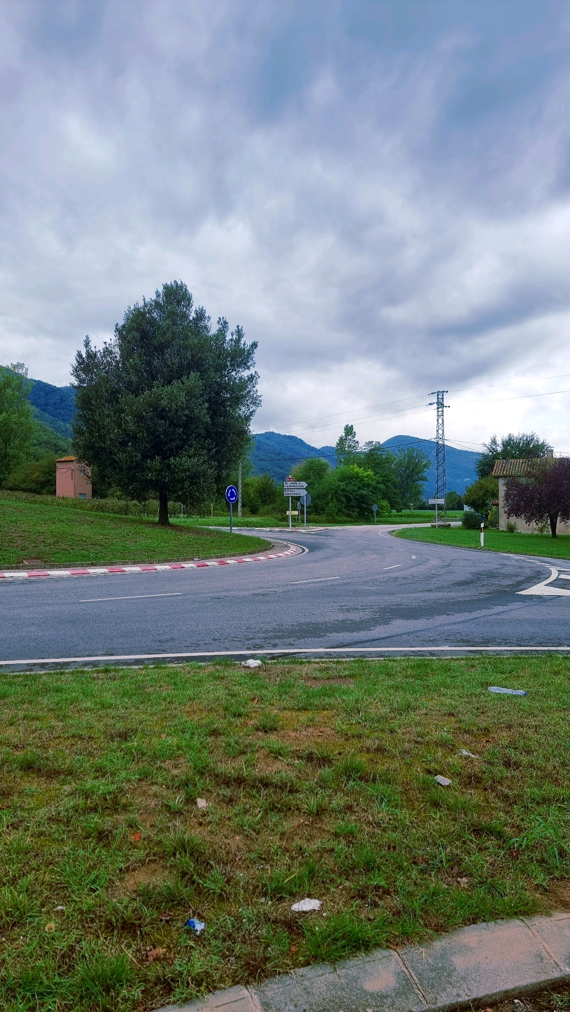 The image depicts an outdoor scene at a road intersection under an overcast sky. In the foreground, a paved road curves to the right. The road has white lane markings and a red and white striped curb on the left side. Beyond the road to the left is a grassy embankment with a lone, mature tree. Further up the embankment, a small, light-pink building is visible. To the right of the road, more grass and a few scattered pieces of litter are present. In the mid-ground, several trees and foliage are visible, along with road signs. One signpost has multiple directional arrows, with text including "CAMPRODON" and "ANDORRA". A blue circular sign with a white arrow indicating a roundabout is also present. A tall electricity pylon stands to the right of the intersection, with a building featuring a red-tiled roof nearby. In the background, rolling hills covered in trees form the landscape, and mountains are visible in the distance under the cloudy sky. The scene is located on the N-260 road near the entrance to the town of La Vall de Bianya, Girona, Spain.