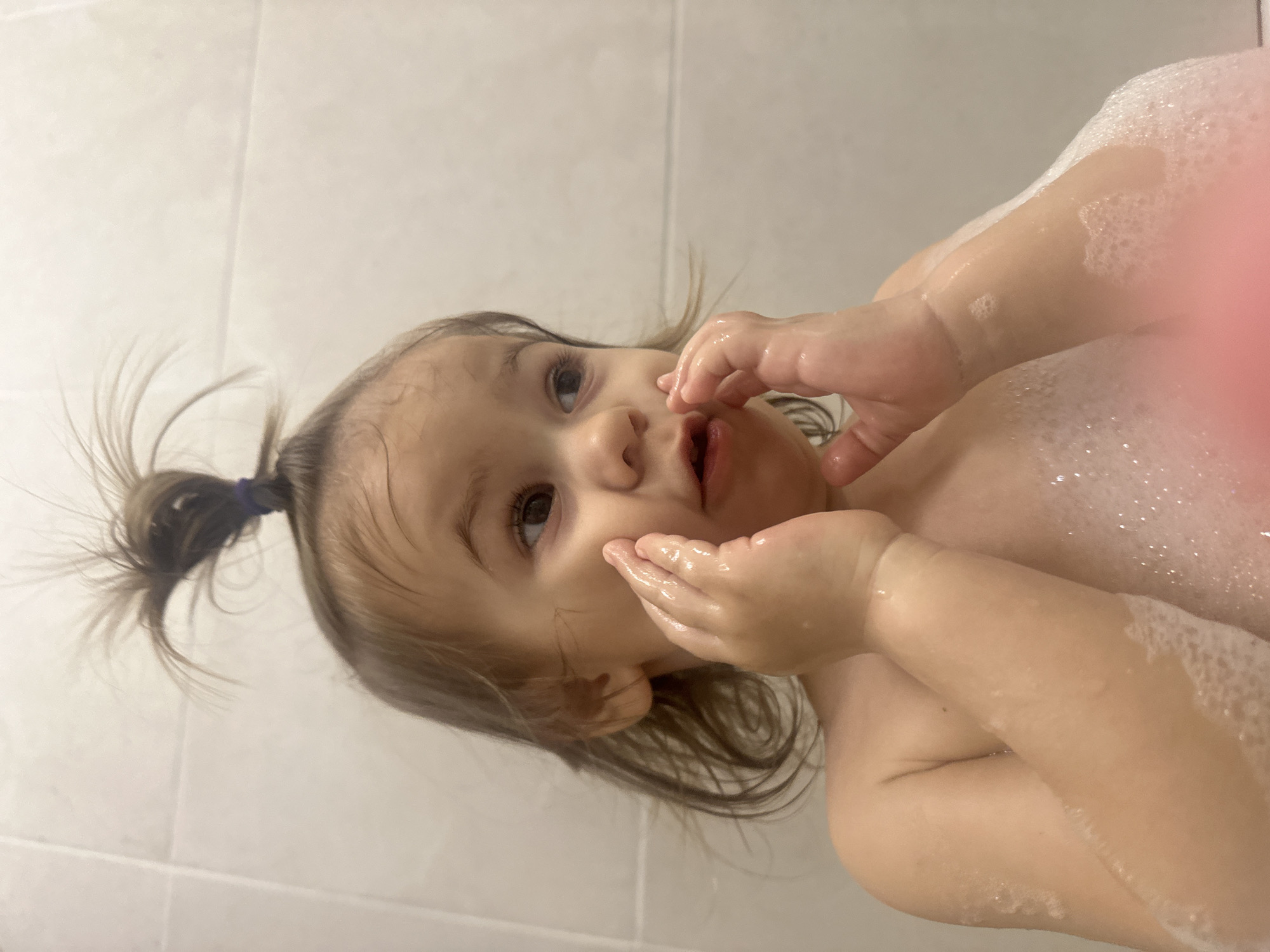 A young child with light skin and brown hair pulled into a small ponytail with a blue hair tie is partially submerged in a bathtub filled with soapy water. The child's hands are near their face, with water droplets visible on their skin. Some body parts, including arms and chest, are covered in white bubbles. The background consists of white tiles, possibly a shower or bathroom wall. No evidence suggests the location as Kharnavy, Russia.