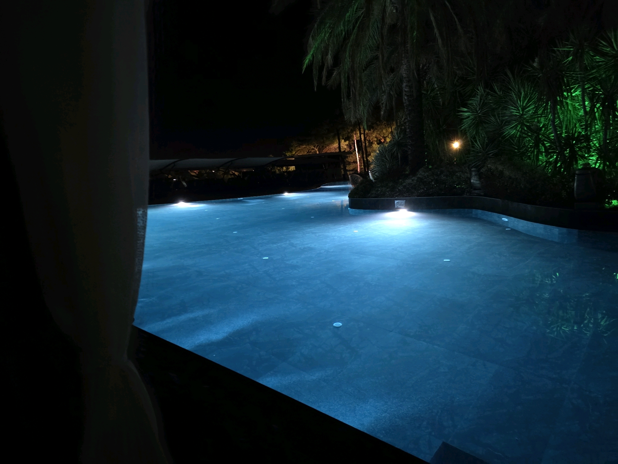 A nighttime view of an illuminated swimming pool at a resort in Bodrum, Turkey. The pool is a deep blue and illuminated by underwater lights. Palm trees and lush green foliage, lit with green lights, surround the pool on the right side. In the background, the roof of a building with lights is visible, along with a hint of trees. The foreground on the left shows a dark, blurred shape that appears to be a curtain or fabric.